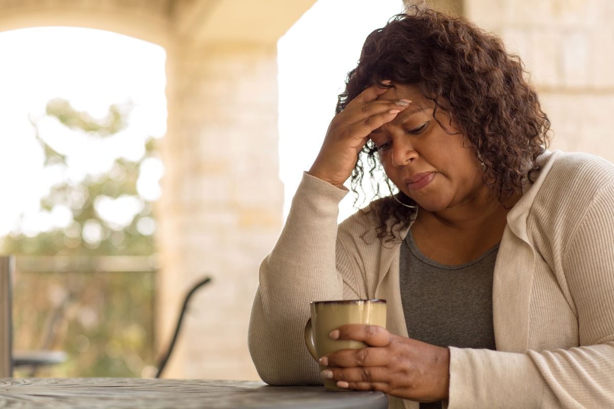 Woman holding a mug and rubbing her forehead at a table, looking tired or stressed.