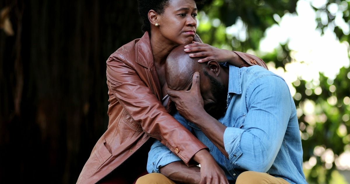 Two people sit close together outdoors, one comforting the other with a hand on their head.