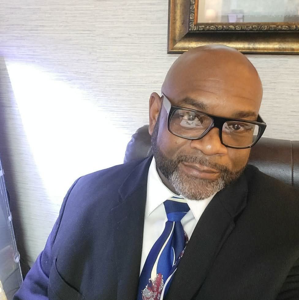 Man in a dark suit and blue tie, wearing glasses, seated indoors and looking at the camera.