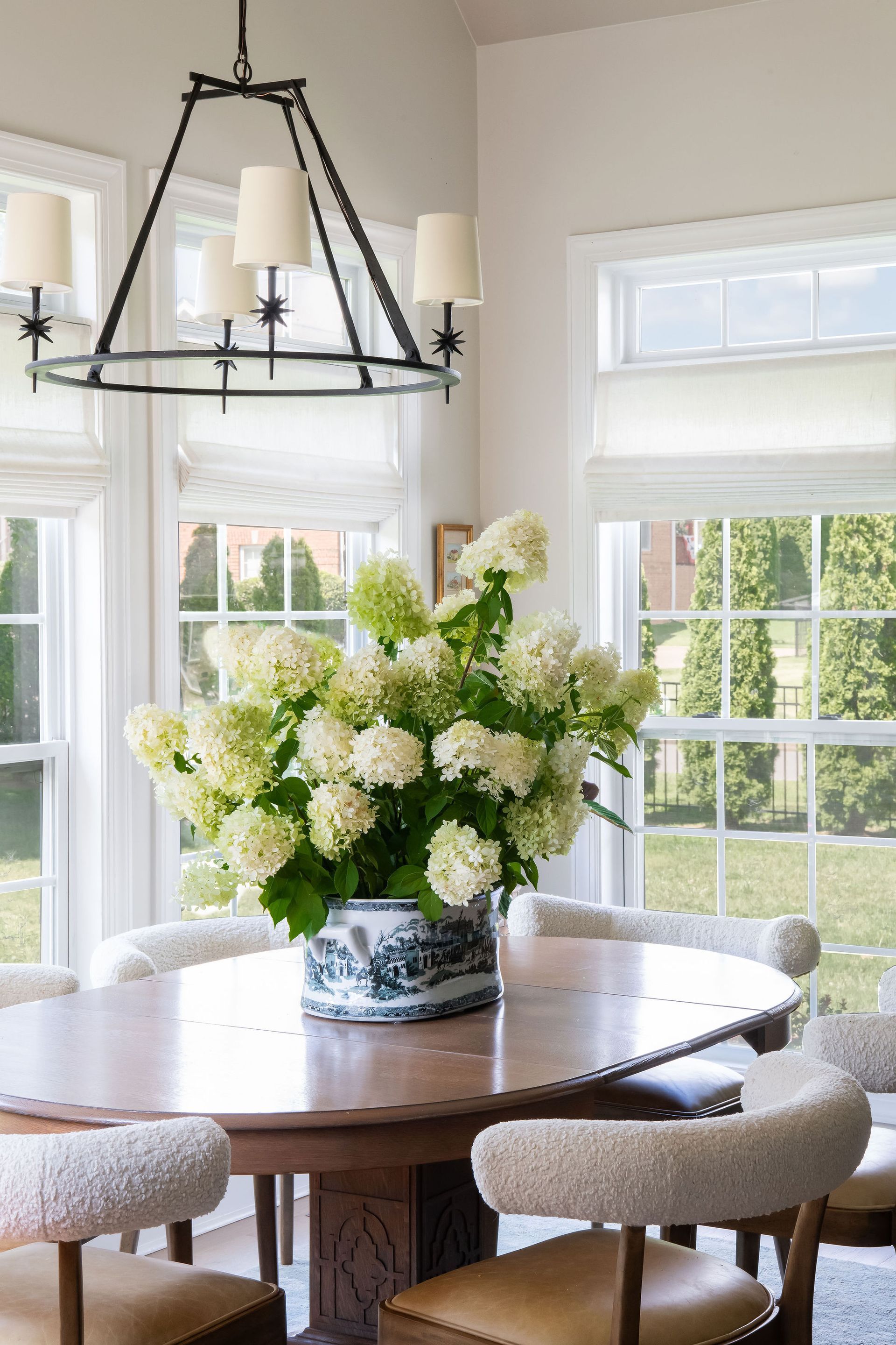 Dining room with round wooden table, white flowers in a vase, and large windows with blinds