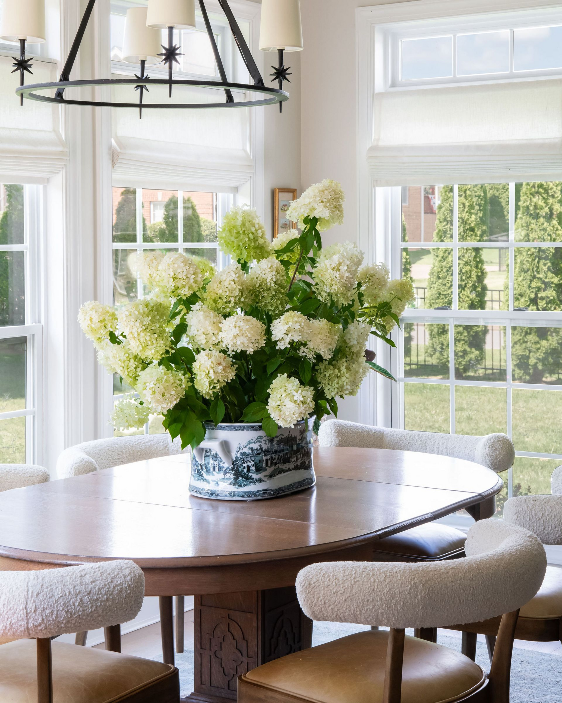 Dining room with round wooden table, white flowers in a vase, and large windows with blinds