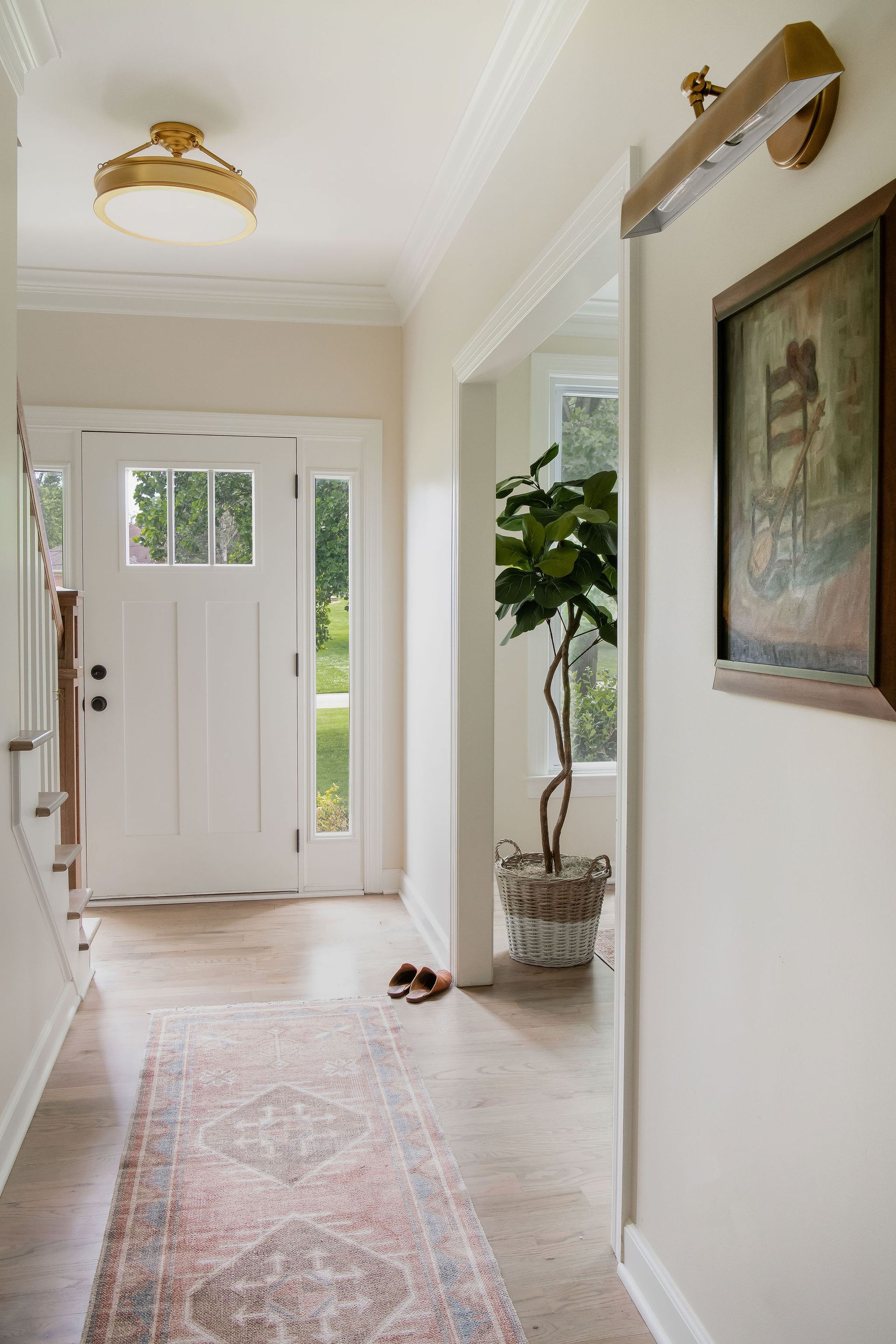 Hallway with white walls, patterned rug, front door, and a painting; a large potted plant is visible through an open doorway.