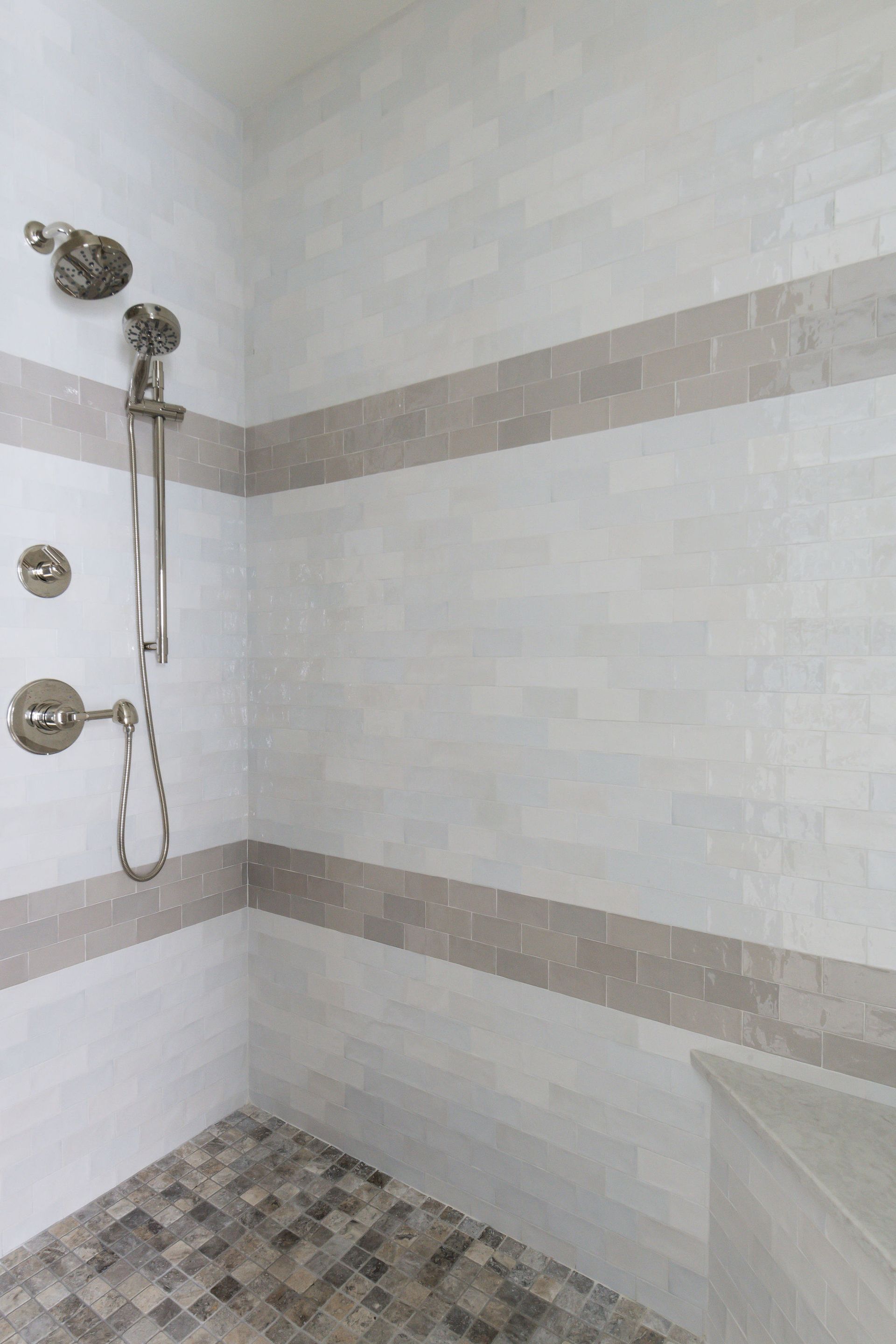 Shower with white and gray tiled walls, stainless steel fixtures, and mosaic floor.