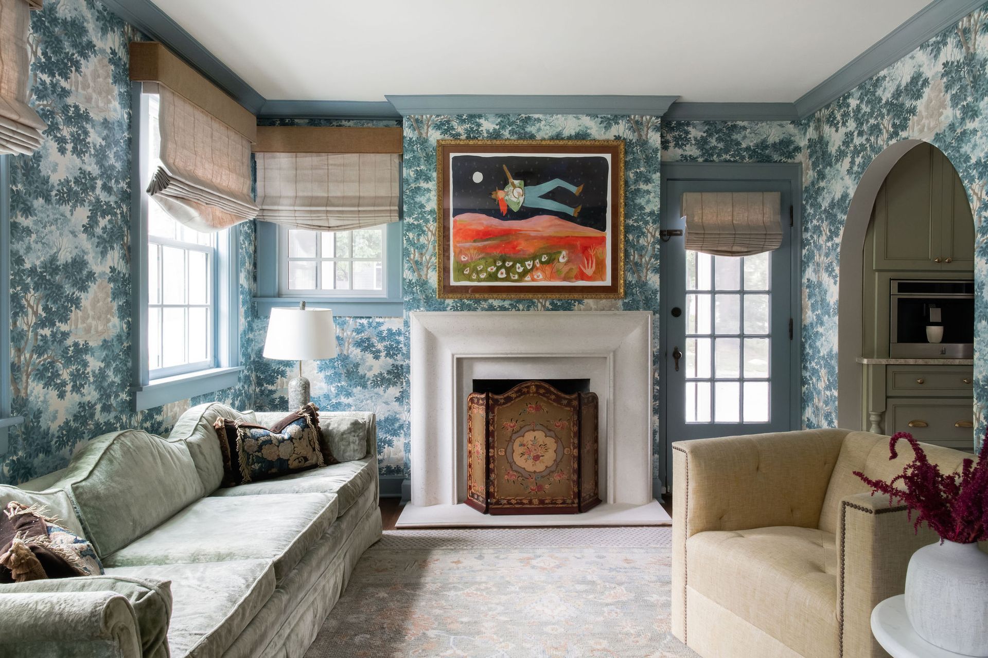 A living room with blue floral wallpaper, a white fireplace, a sofa, and an armchair under a painting of a figure.
