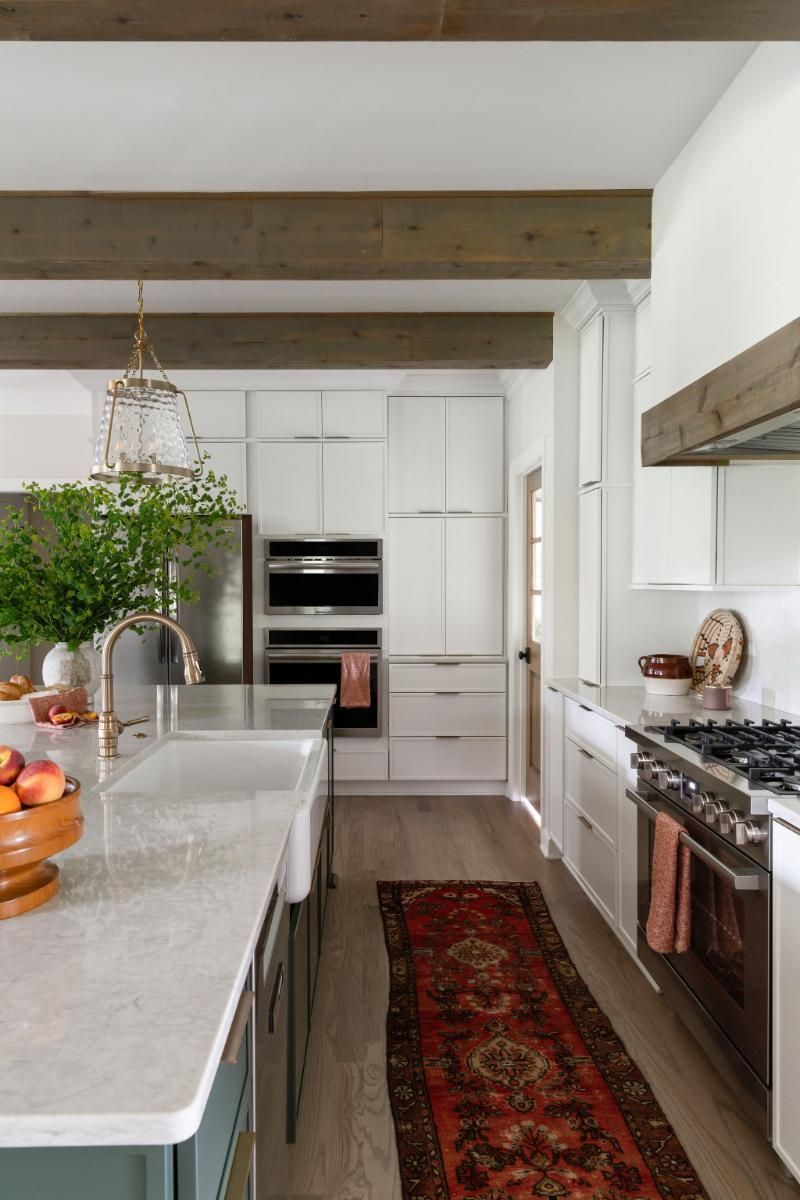 Bright kitchen with white cabinetry, stone island, exposed beams, and red runner rug