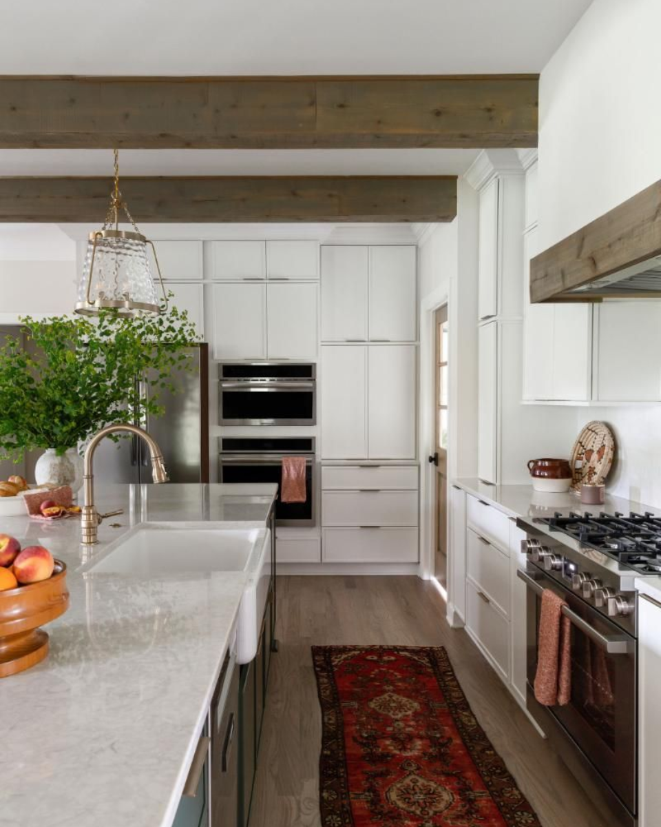Bright kitchen with white cabinetry, stone island, exposed beams, and red runner rug