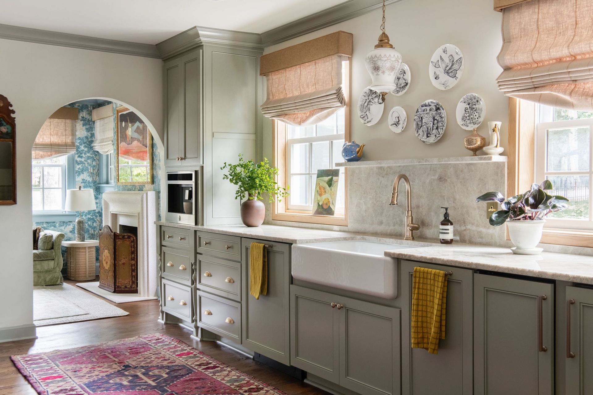 Sage green kitchen with a farmhouse sink, floral rug, and plates on the wall.