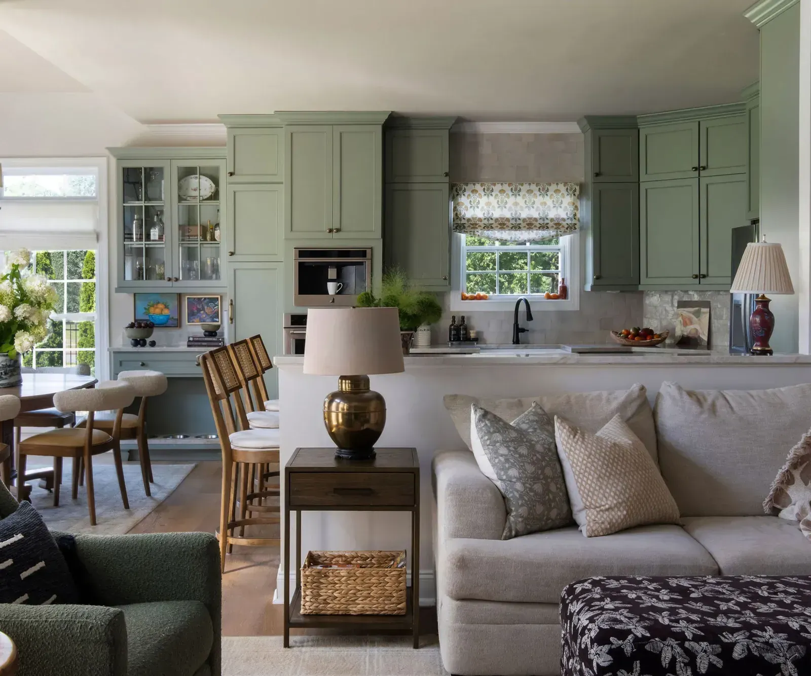 Open-concept living room featuring a small table lamp on a side table next to a sofa, looking into a kitchen with sage green cabinetry and bar seating.