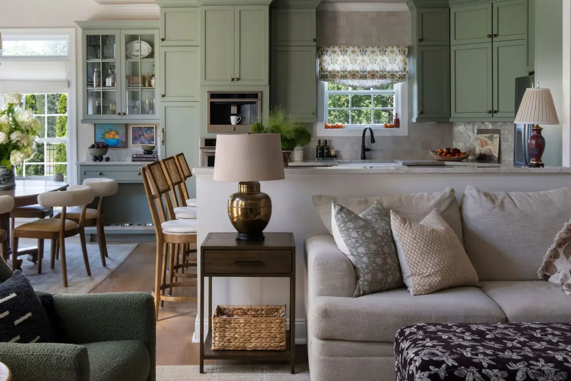 Open-concept living room featuring a small table lamp on a side table next to a sofa, looking into a kitchen with sage green cabinetry and bar seating.