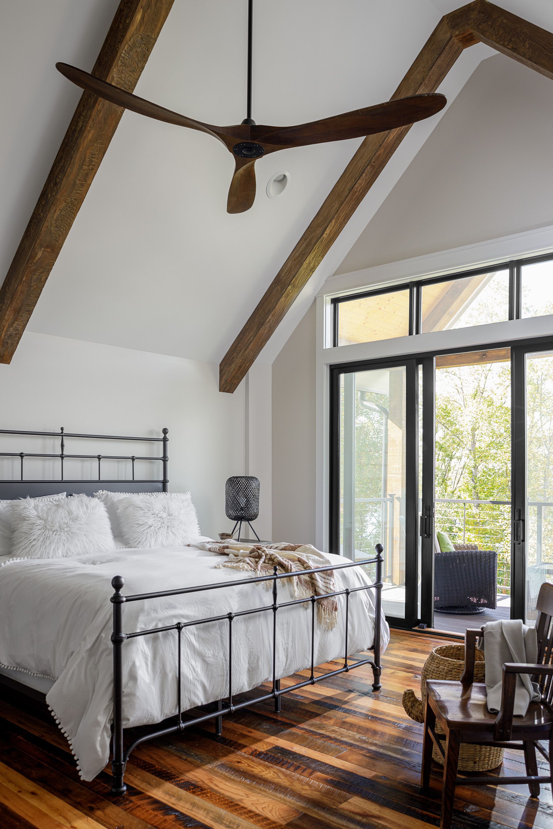 Bright bedroom with vaulted beams, black metal bed, striped rug, and glass doors to greenery