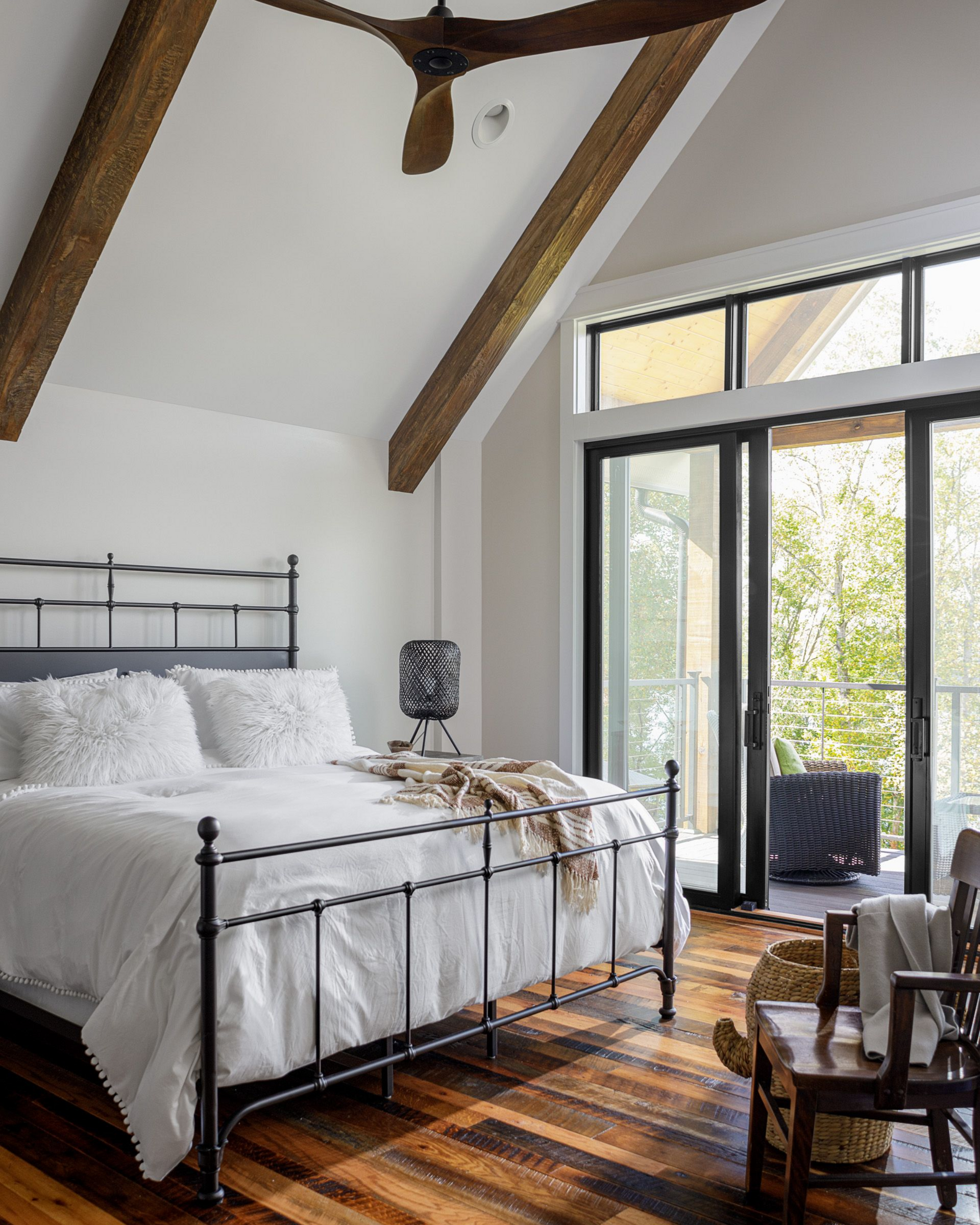 Bright bedroom with vaulted beams, black metal bed, striped rug, and glass doors to greenery