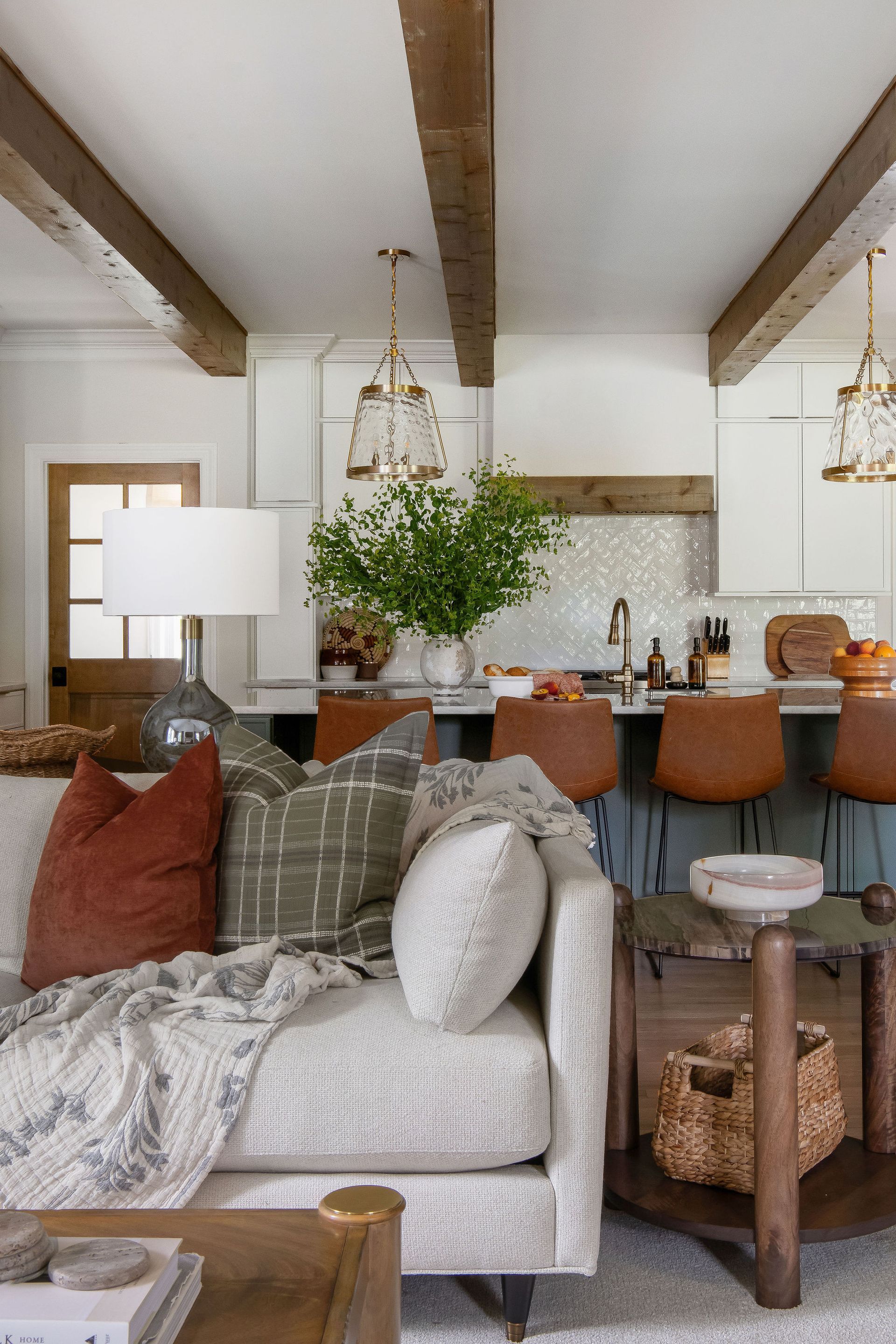 Cozy living room with a cream sofa, rust and green pillows, and exposed wooden beams above a kitchen bar.