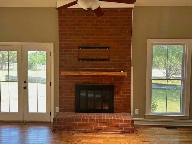 Brick fireplace with wooden mantel, flanked by white-framed doors and window; brown walls, hardwood floor.