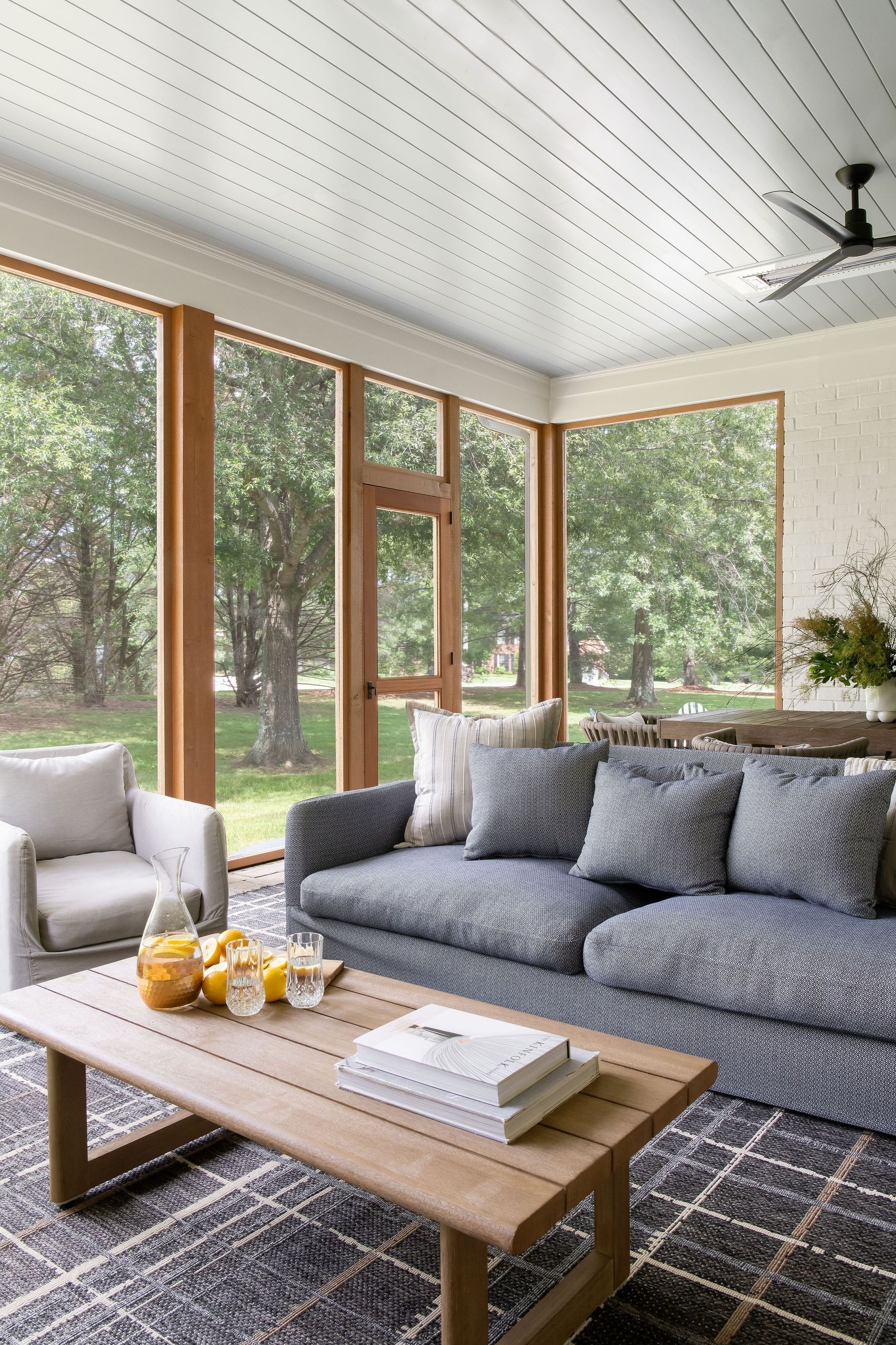 Sunroom with blue couch, wooden coffee table, and large windows looking onto a lush green yard.