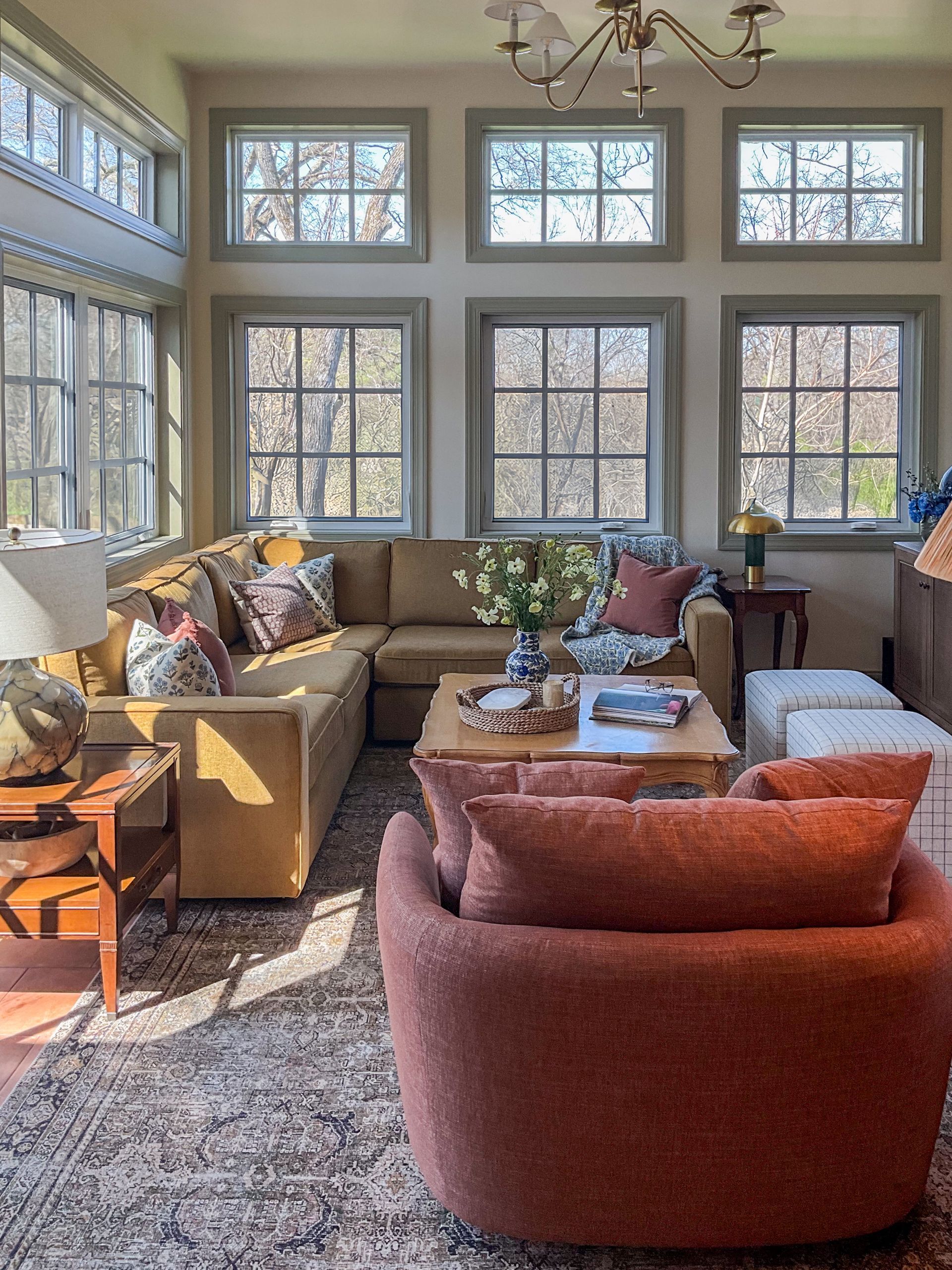 Cozy living room with mustard sofa, rust armchair, large windows, neutral walls, and floral accents.
