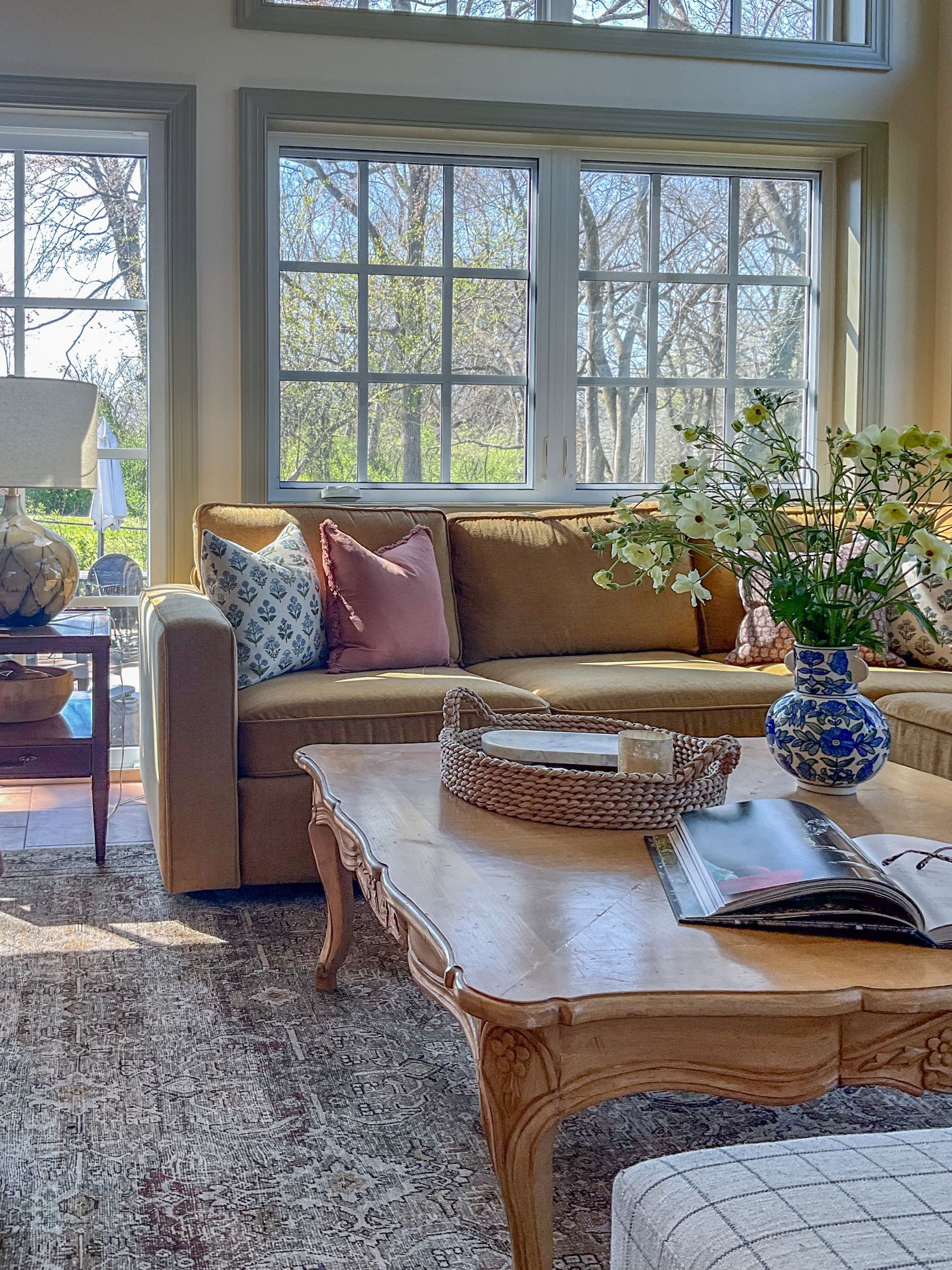 Cozy living room: tan sofa with colorful pillows, floral arrangement, wooden coffee table, and large windows.