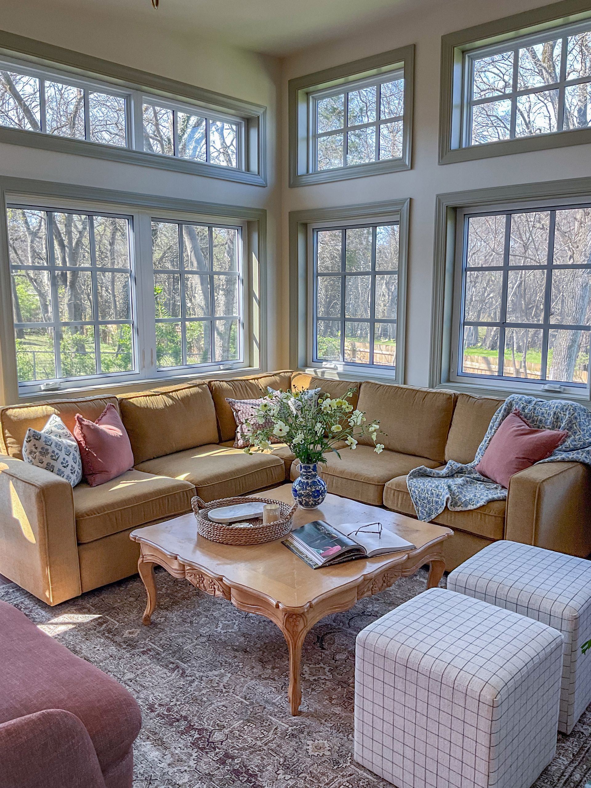 Cozy sunroom with corner sofa, coffee table, ottomans, and large windows overlooking trees. Earth-tone color scheme.