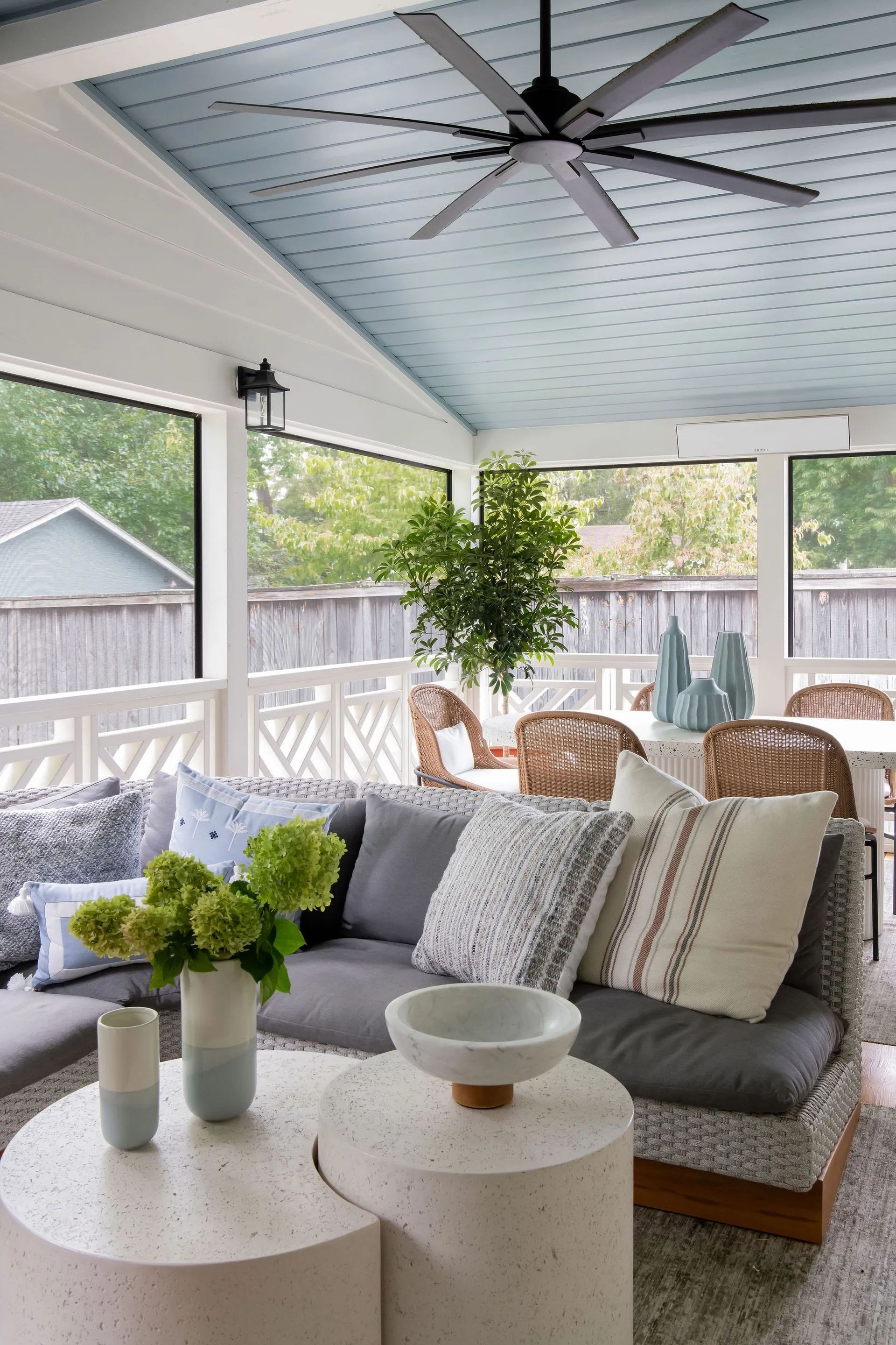 Covered porch with gray sofa, round tables, potted plants, wicker chairs, and a ceiling fan