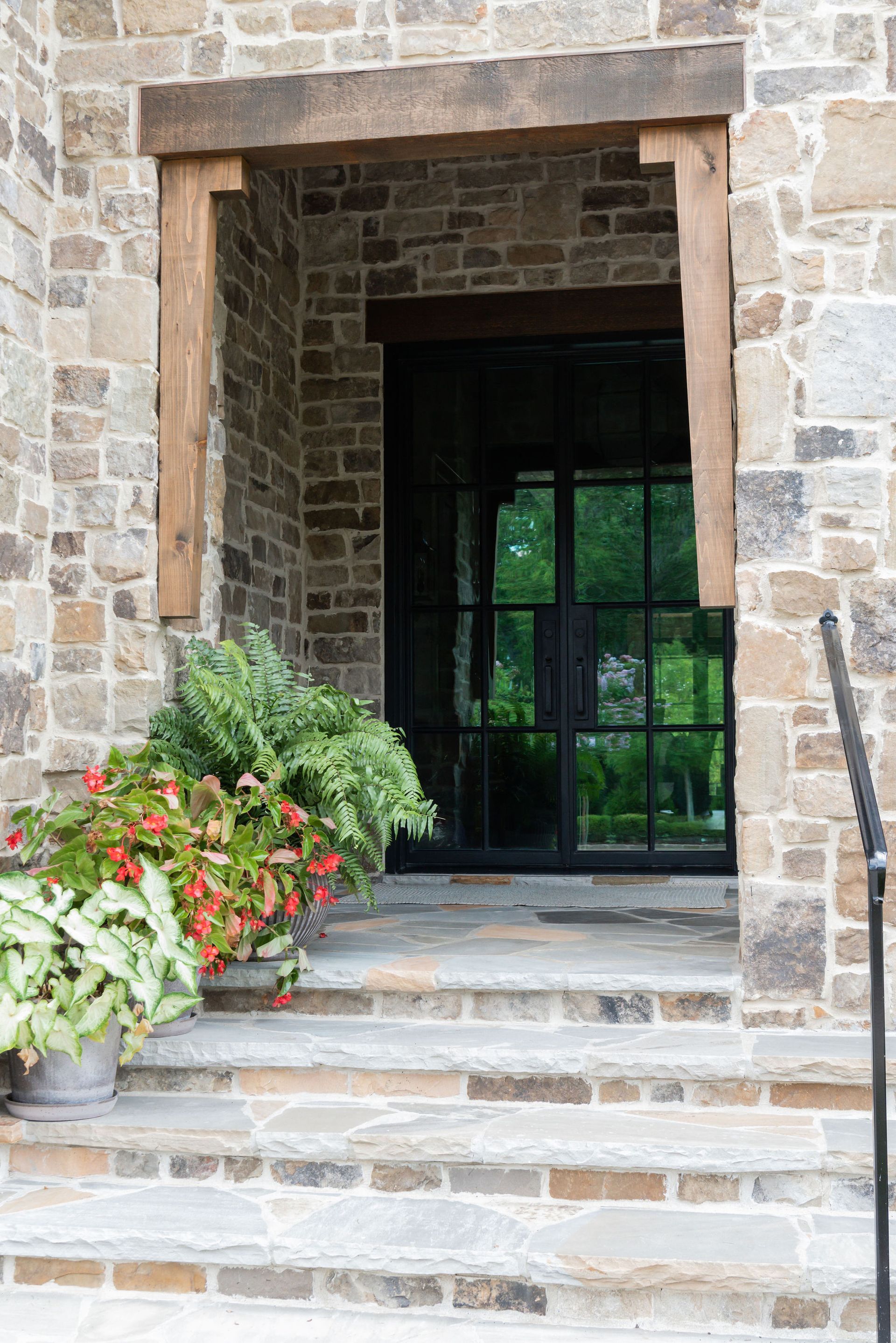 Stone facade entrance with steps, a black metal door, and wooden trim, flanked by potted plants.