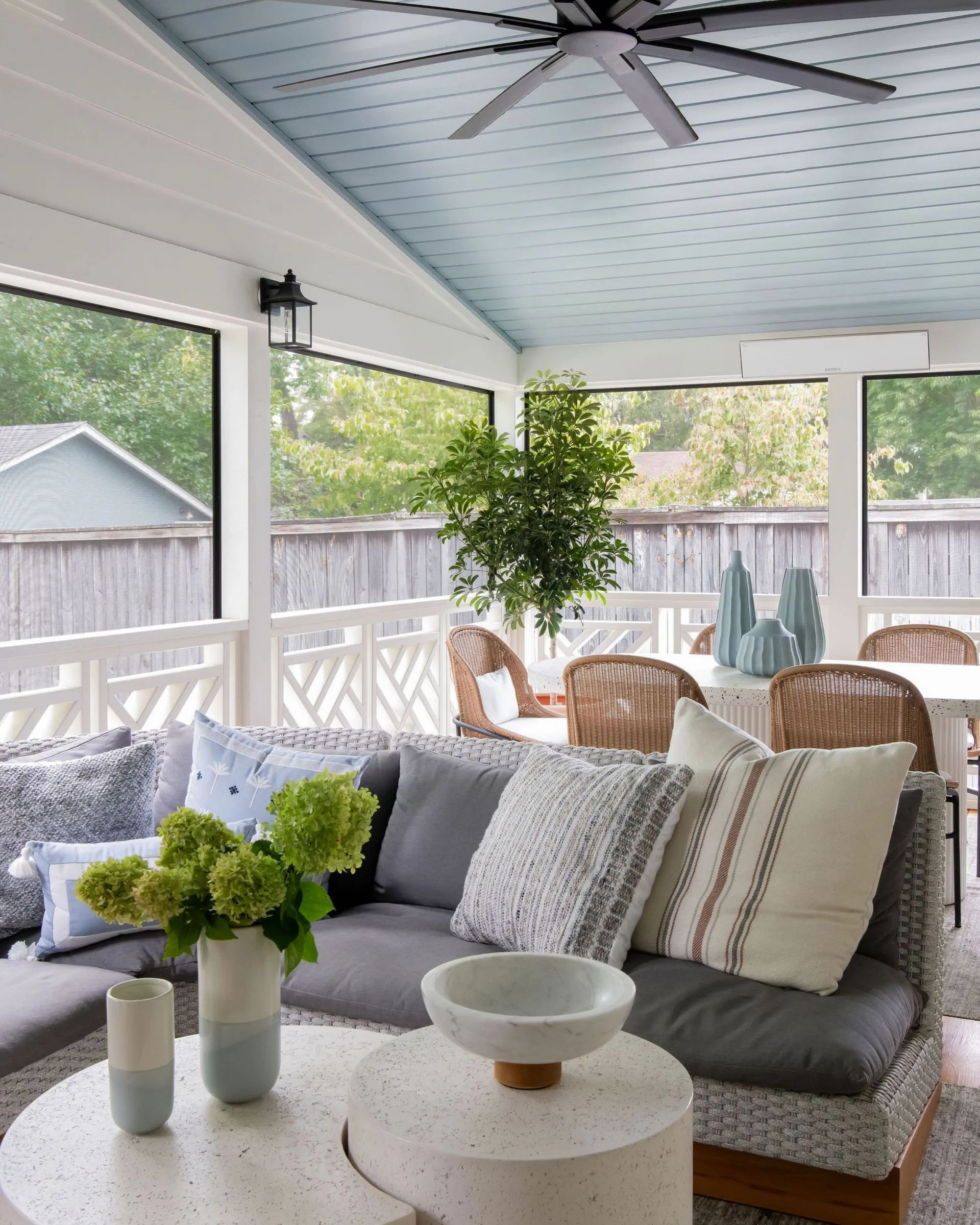 Covered porch with gray sofa, round tables, potted plants, wicker chairs, and a ceiling fan