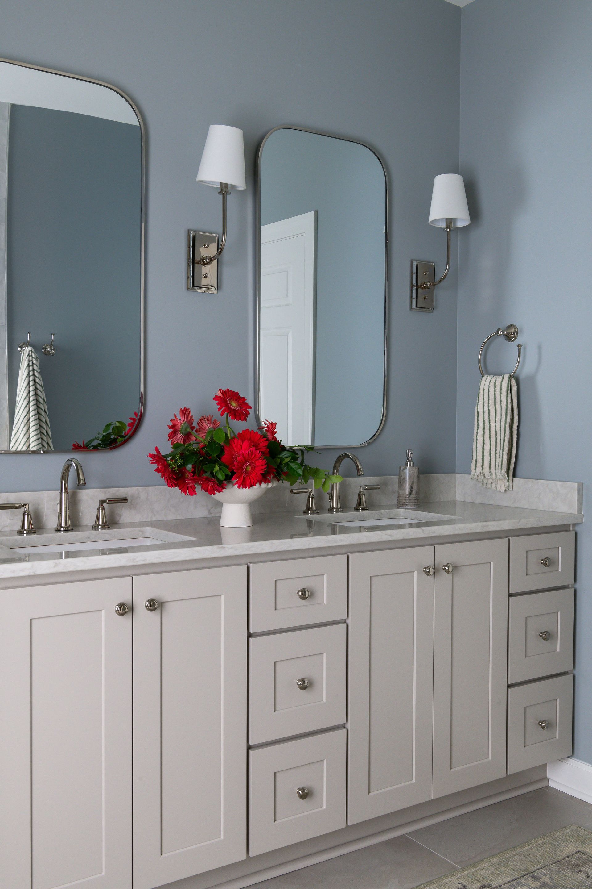 Bathroom with two mirrors, gray cabinets, blue walls, white countertop, and red flowers.