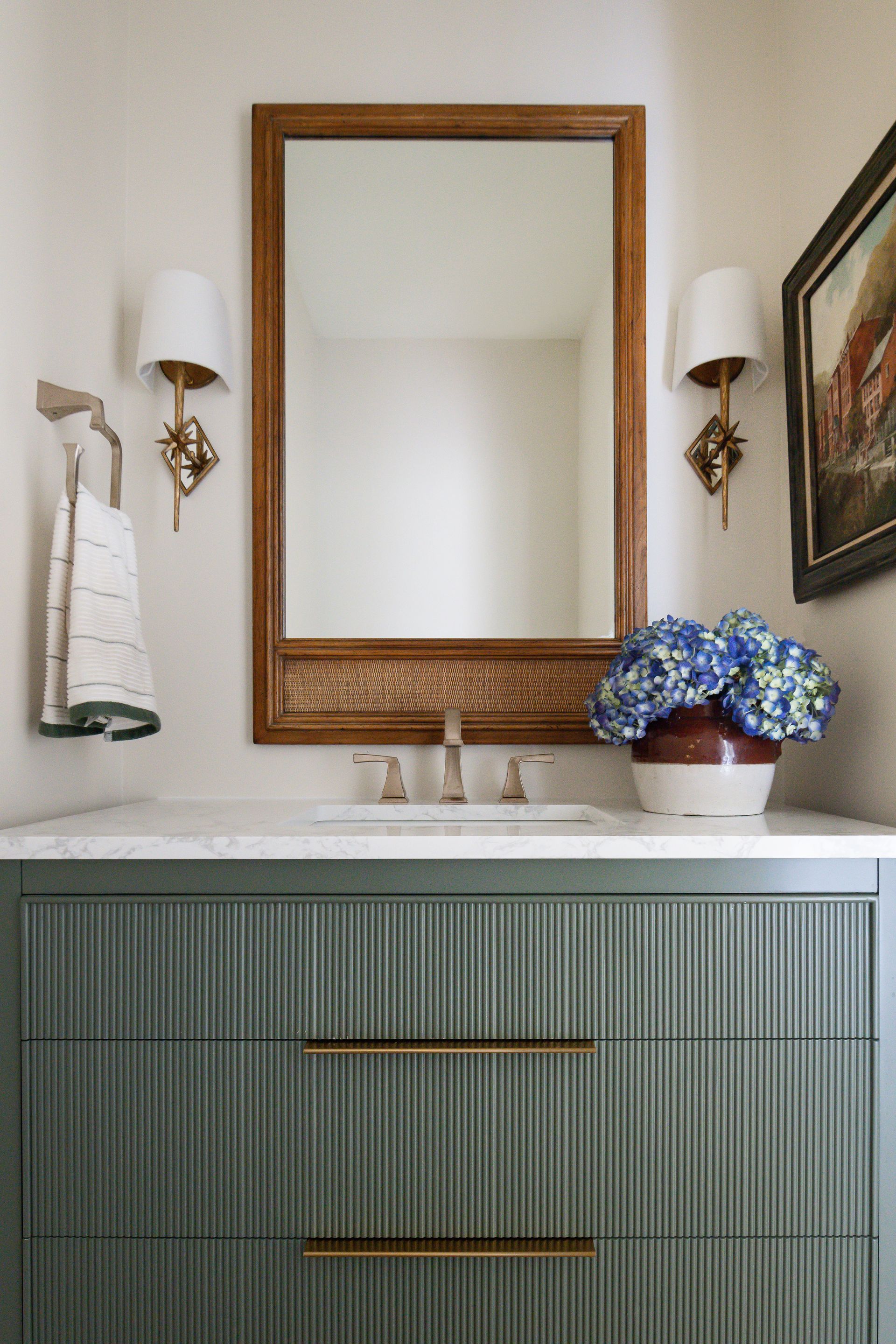 Green bathroom vanity with gold accents, mirror, and blue flower arrangement.