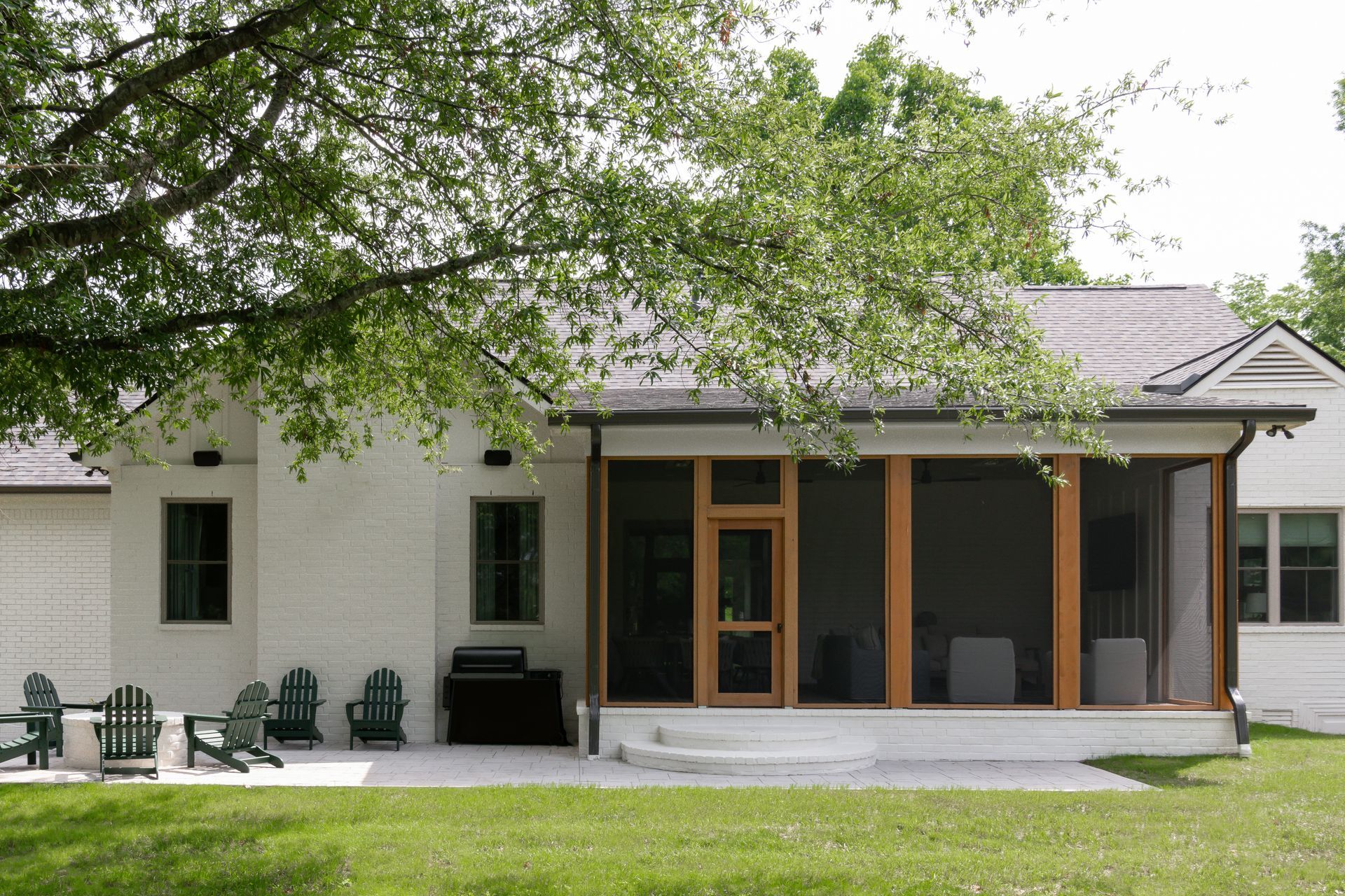 White brick house with a screened porch, patio, and green Adirondack chairs under a tree.