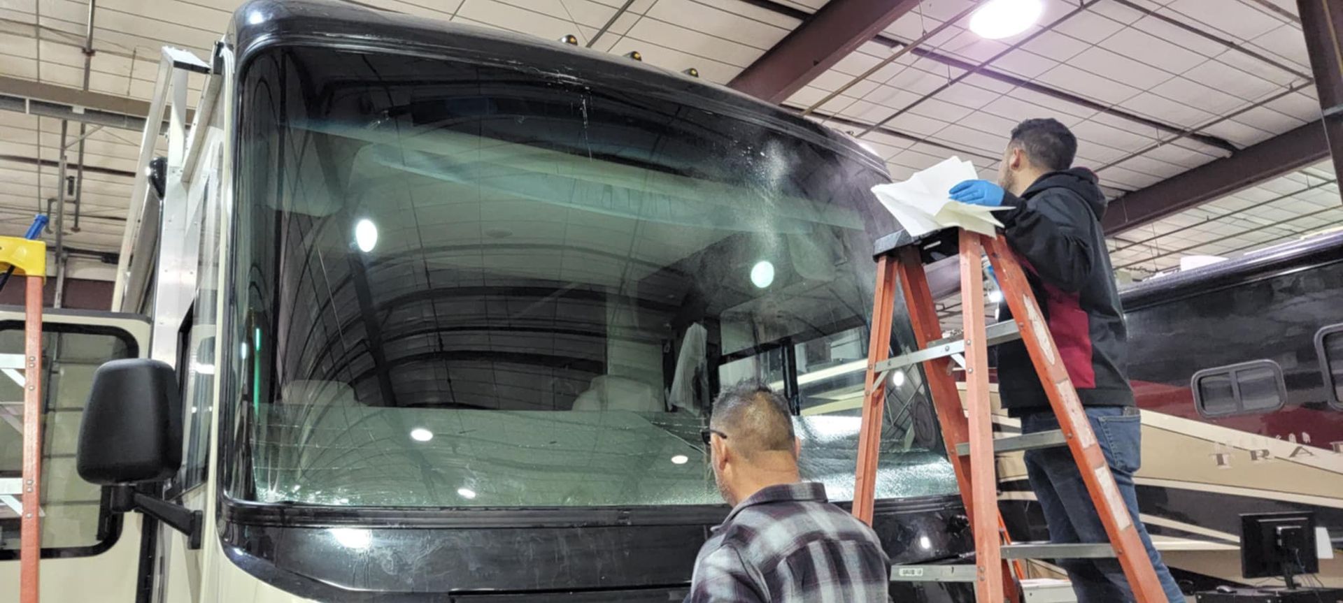 Two workers inspect a bus windshield in a garage, one on a ladder and one standing below.