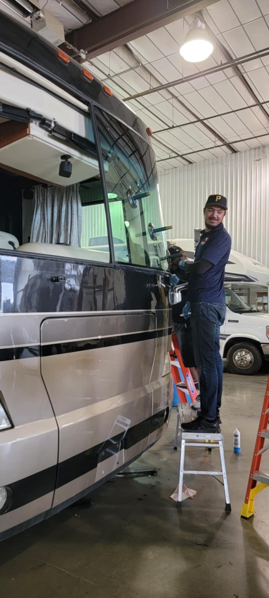 Mechanic on a step stool cleaning the windshield of a beige coach bus in a garage