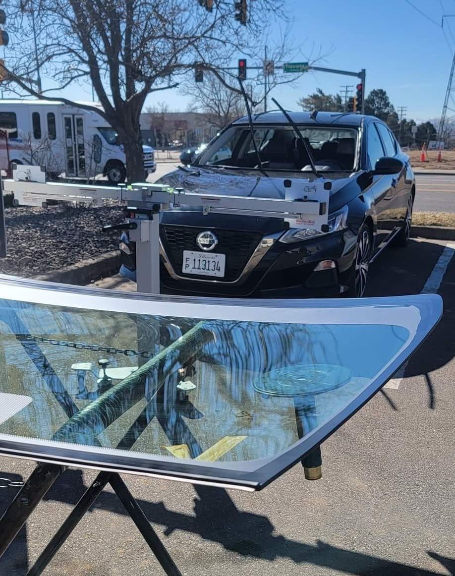 Broken car windshield in front of a black SUV in a parking lot, with a broken glass panel on a stand