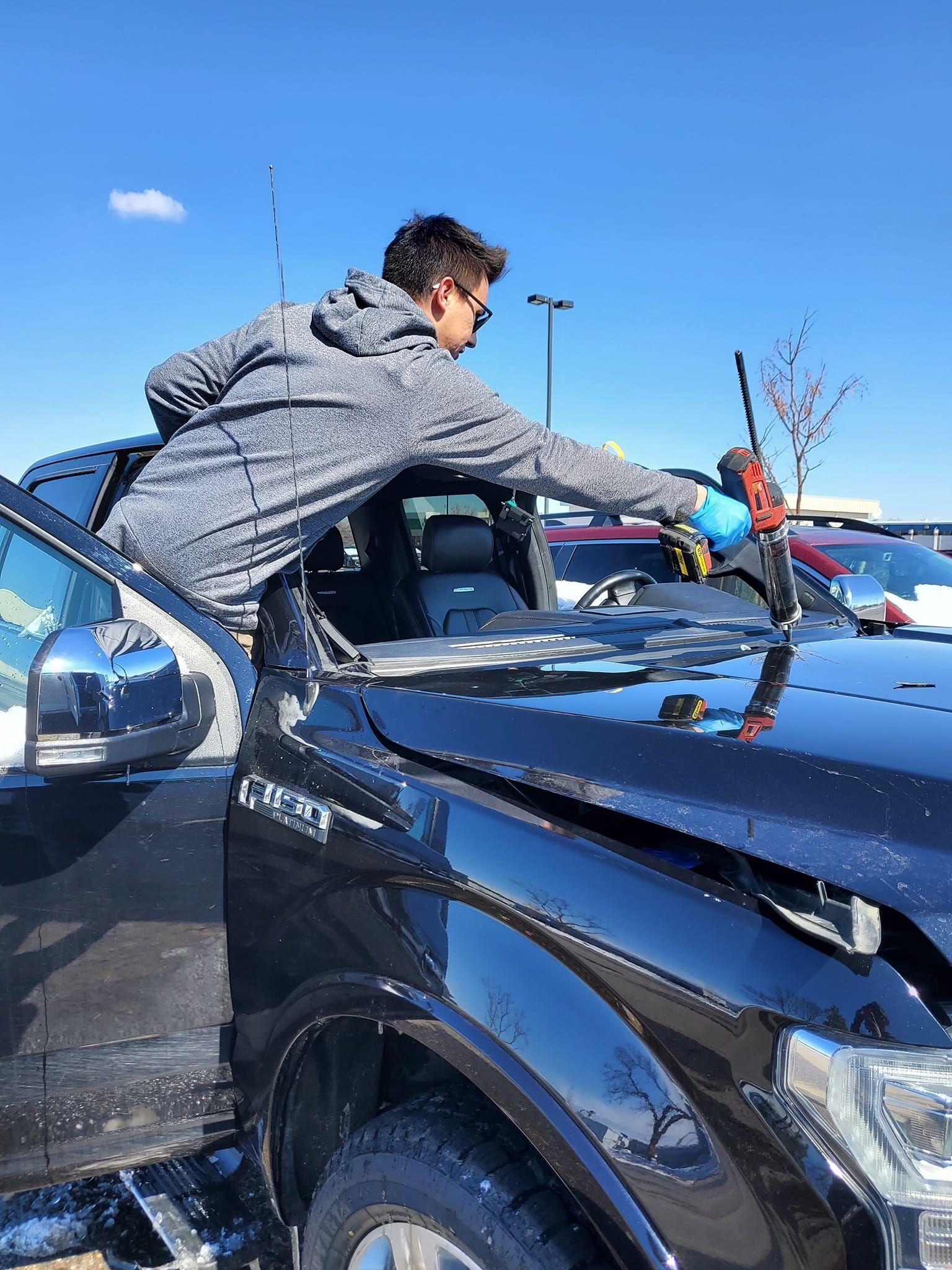 Person reaches over a black car's windshield in a parking lot on a sunny day