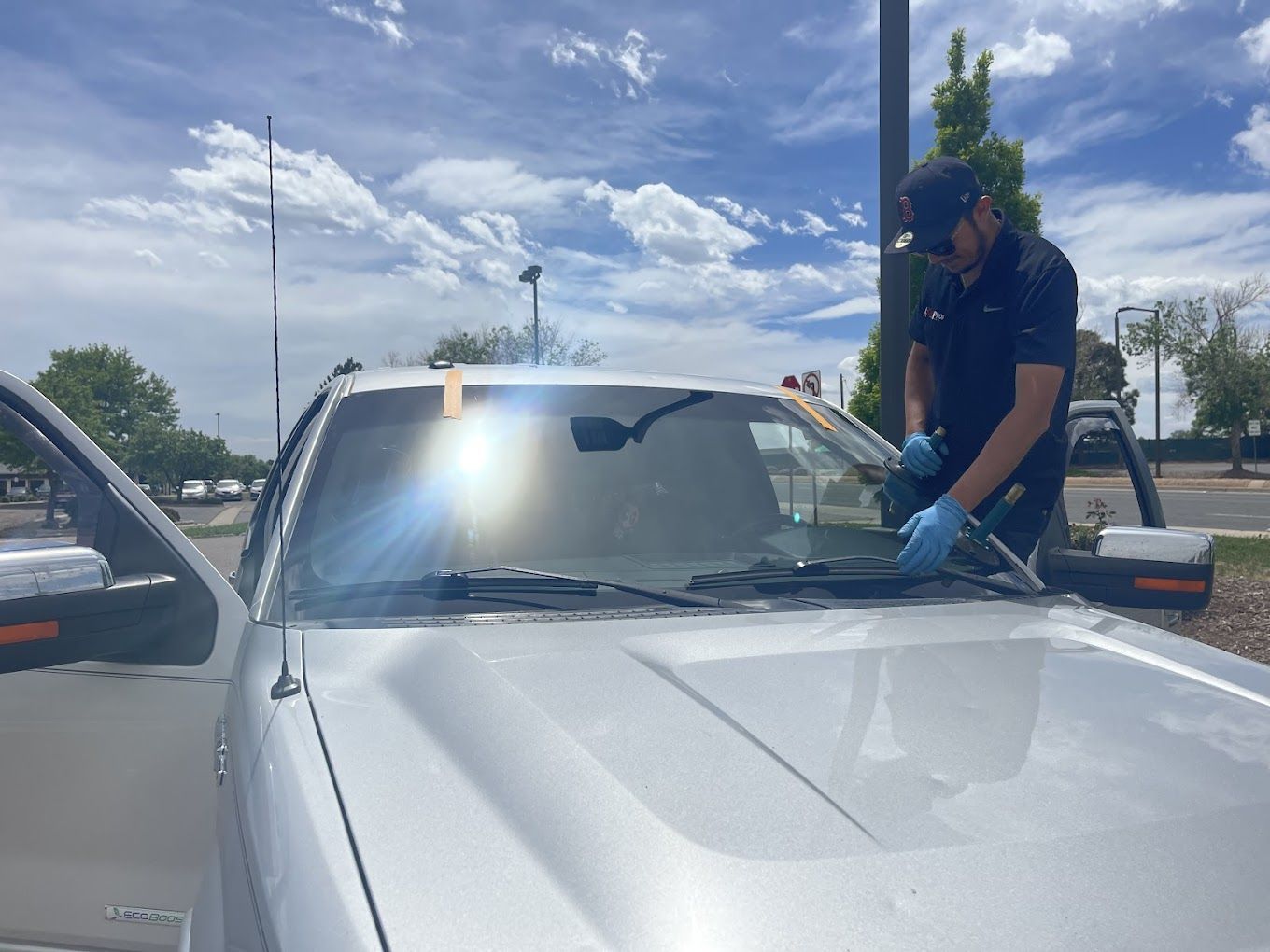 Person cleaning a silver car windshield in a parking lot on a sunny day