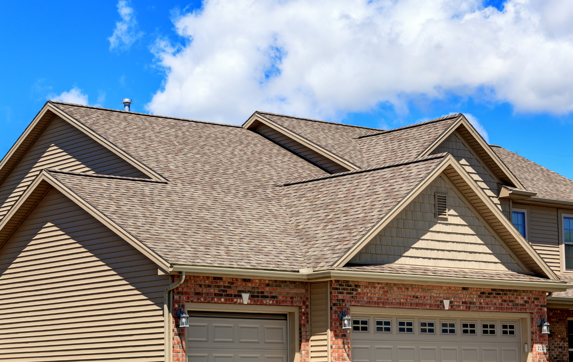 The roof and front exterior of a tan-sided home with a brick-faced garage under a blue sky with scattered clouds.
