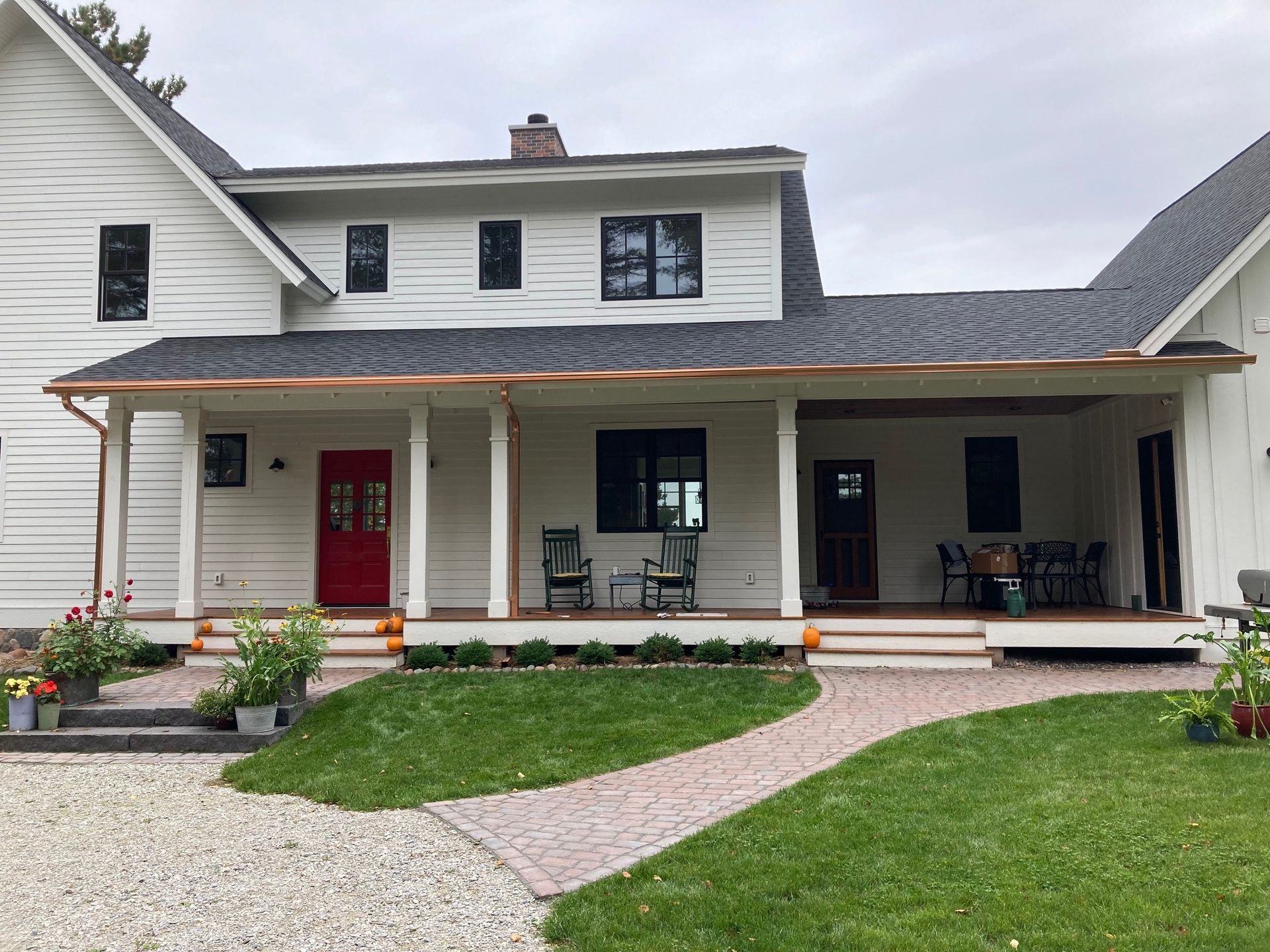 A white farmhouse with a large front porch, black-framed windows, a red front door, and a stone walkway.