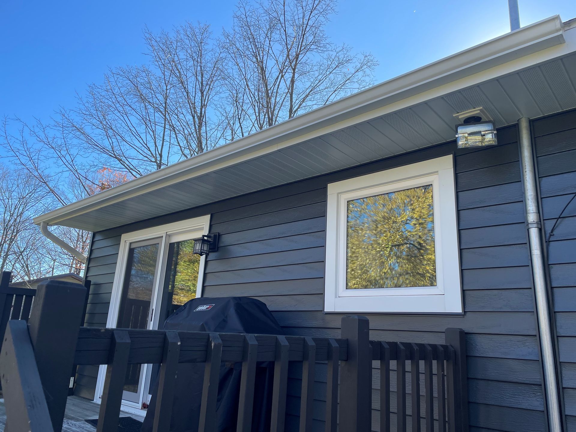 A low-angle view of a dark horizontal-sided house with a white-framed window, sliding glass door, and a wooden deck railing.
