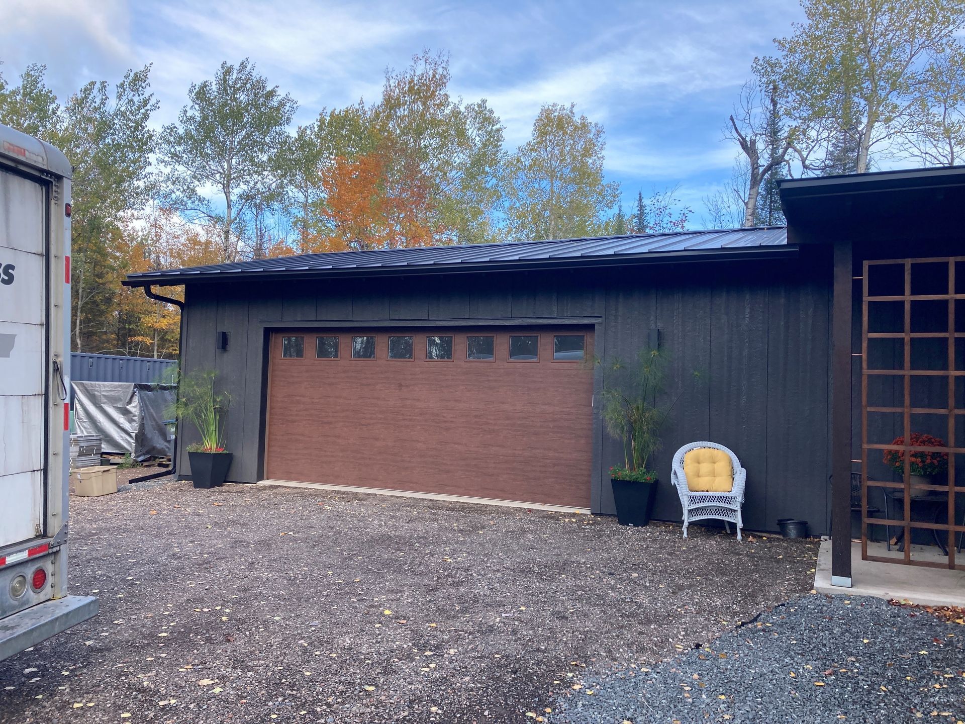 Dark wood garage with a brown door, potted plants, and a wicker chair on a gravel driveway in front of autumn trees.