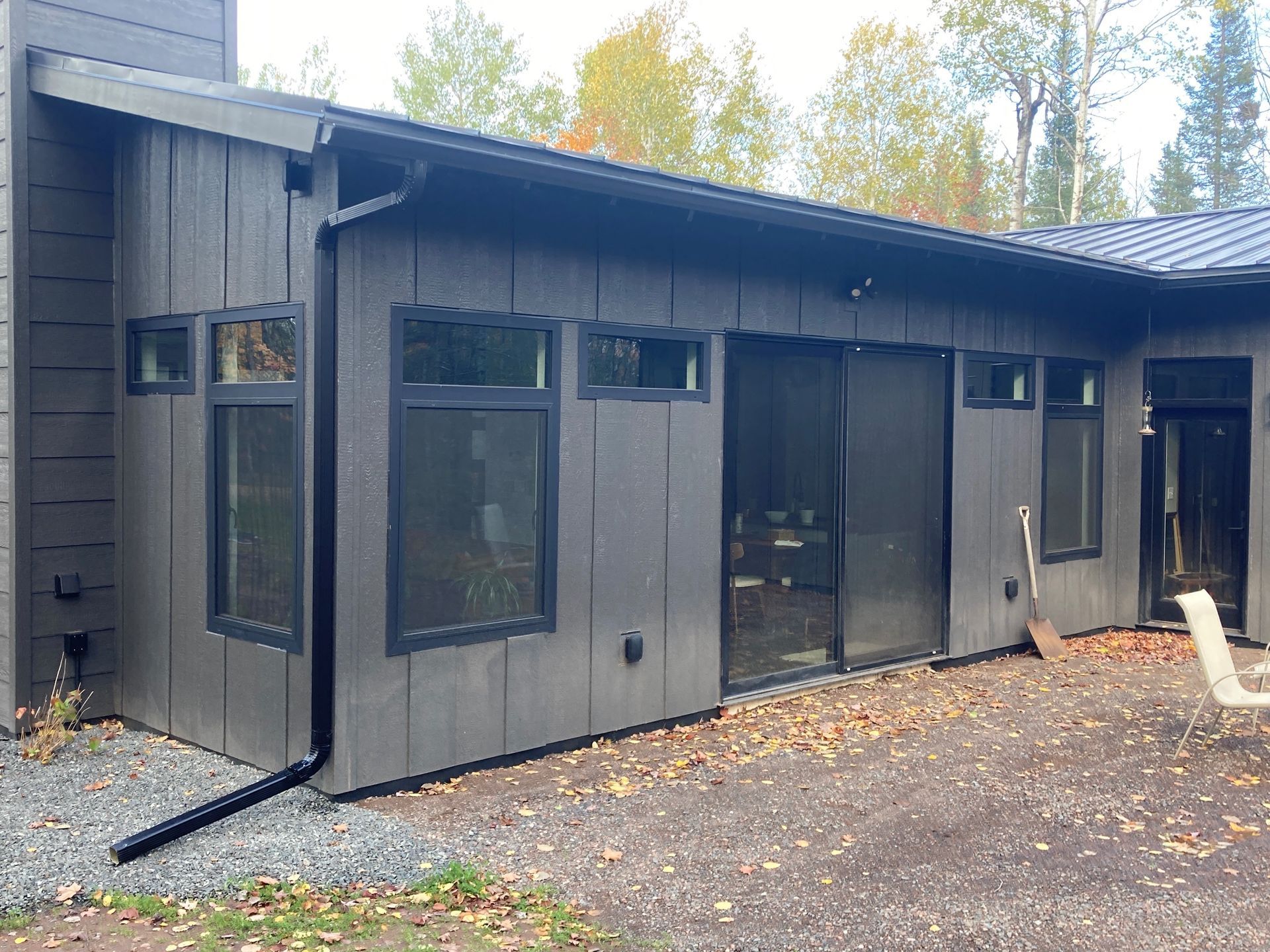 Modern dark gray house with vertical siding, large windows, a black downspout, and a patio with a chair and fallen leaves.