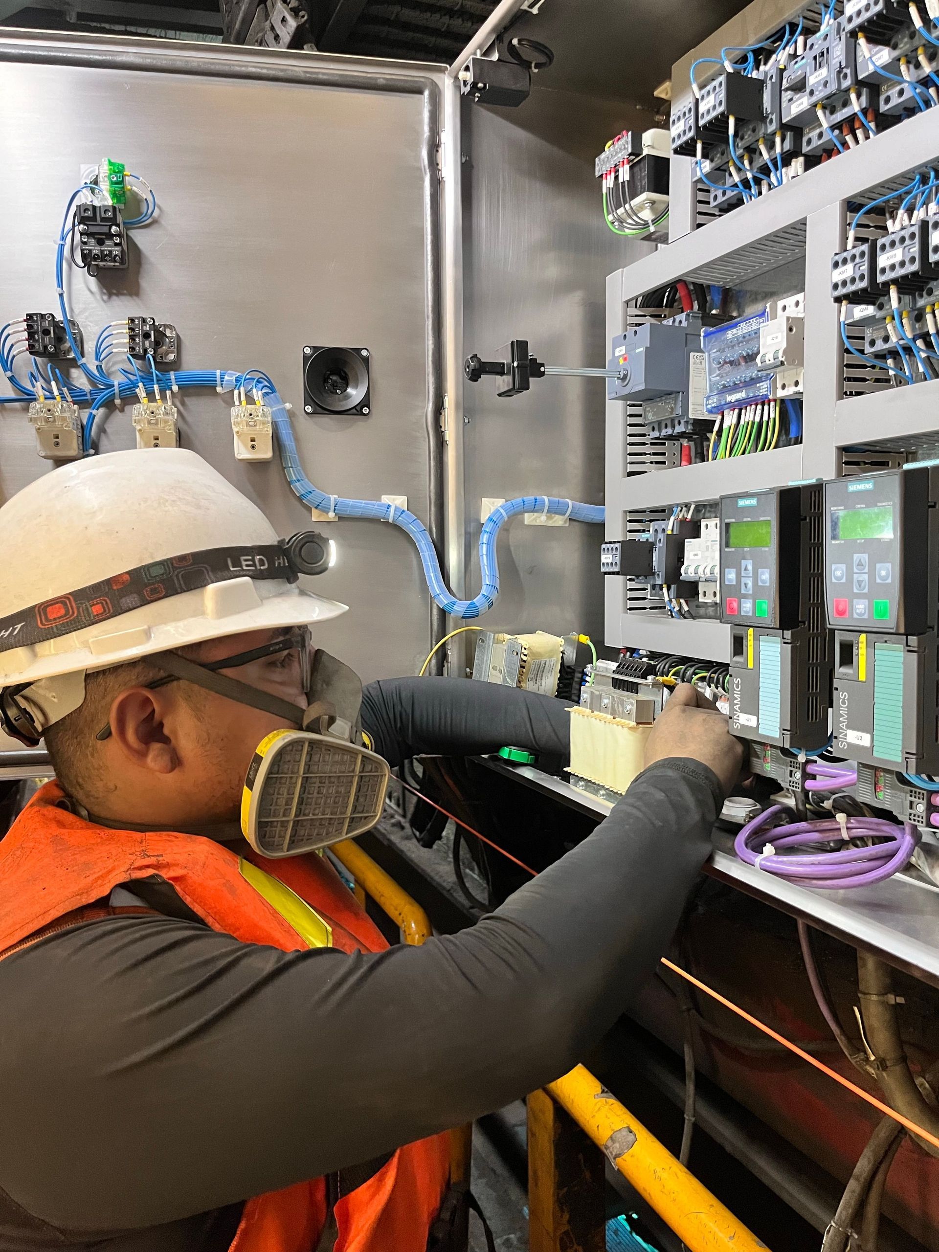 Worker in hard hat and respirator wiring an electrical control panel in a utility cabinet.