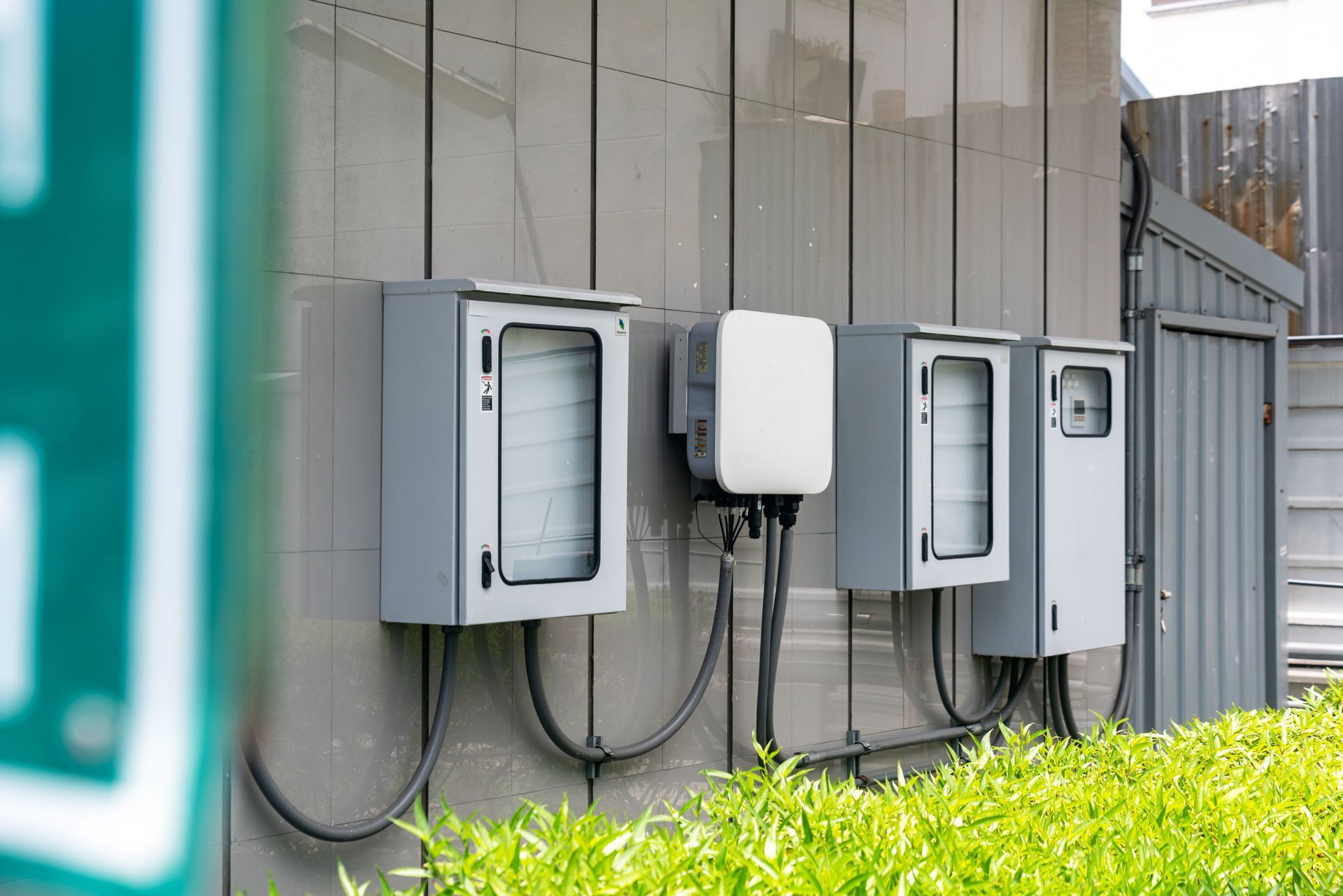 Electrical boxes and cables mounted on an exterior gray wall beside green shrubs.