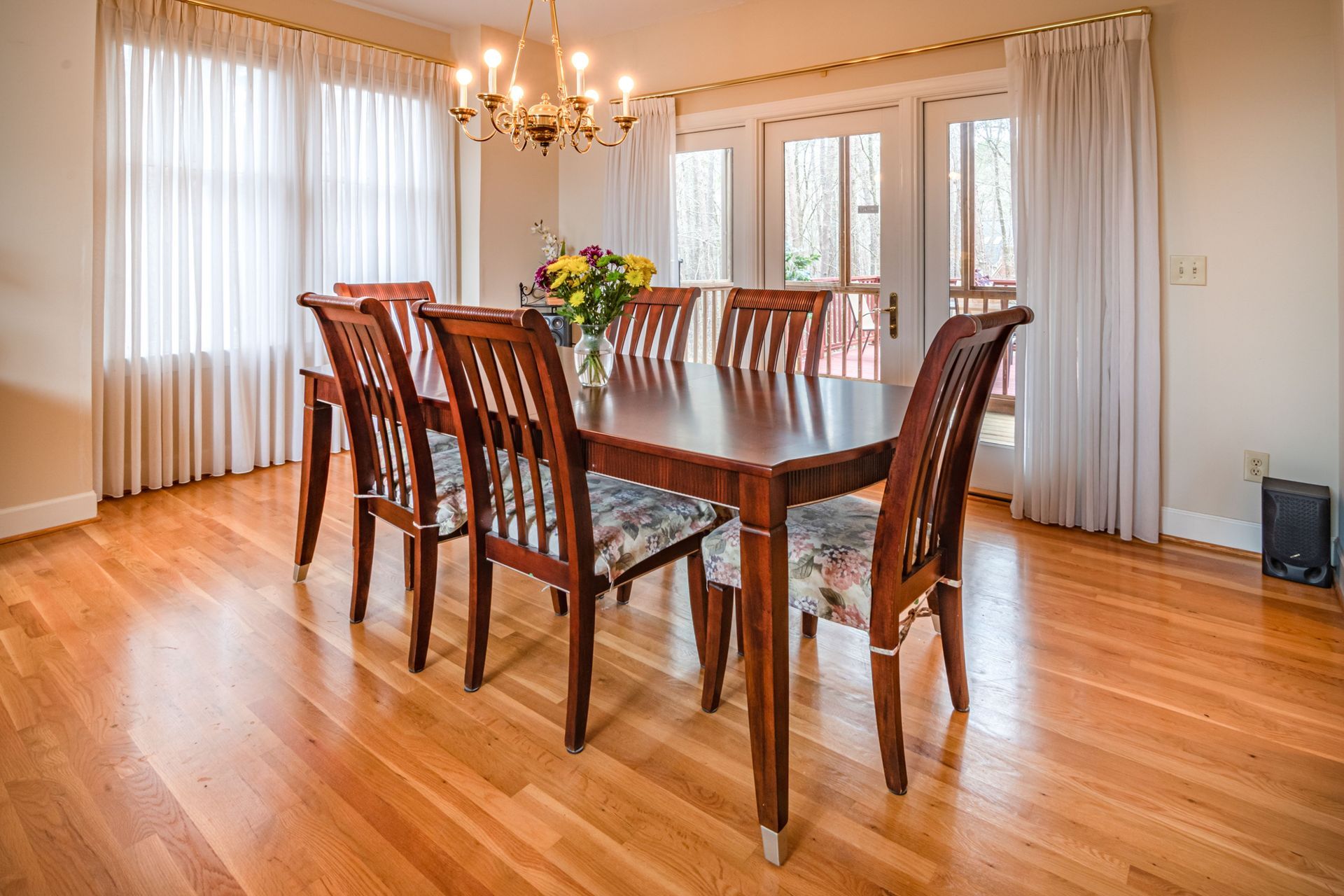 Bright dining room with a long wooden table, matching chairs, chandelier, and white curtains.