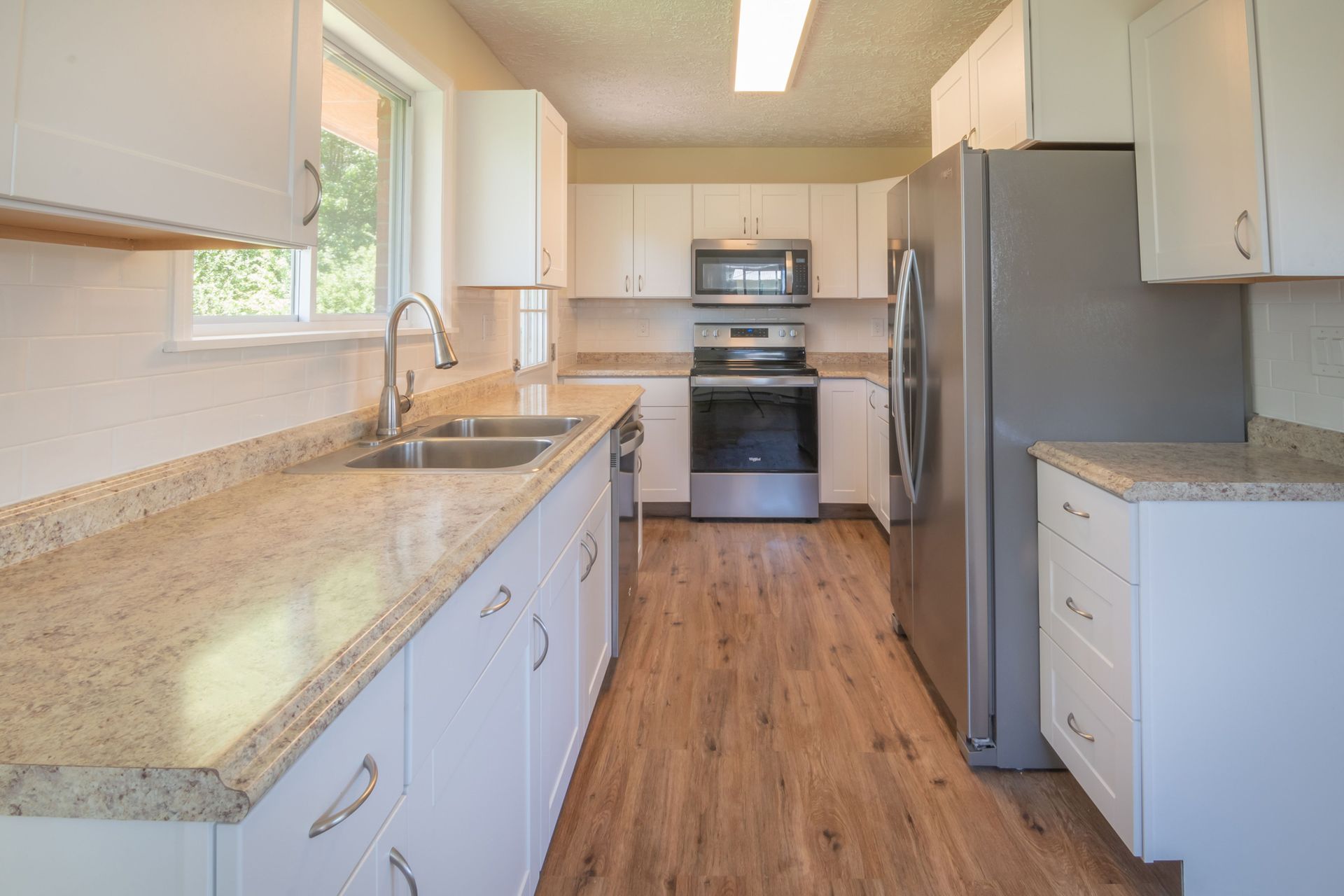 Bright, empty kitchen with white cabinets, granite counters, stainless appliances, and wood floors.