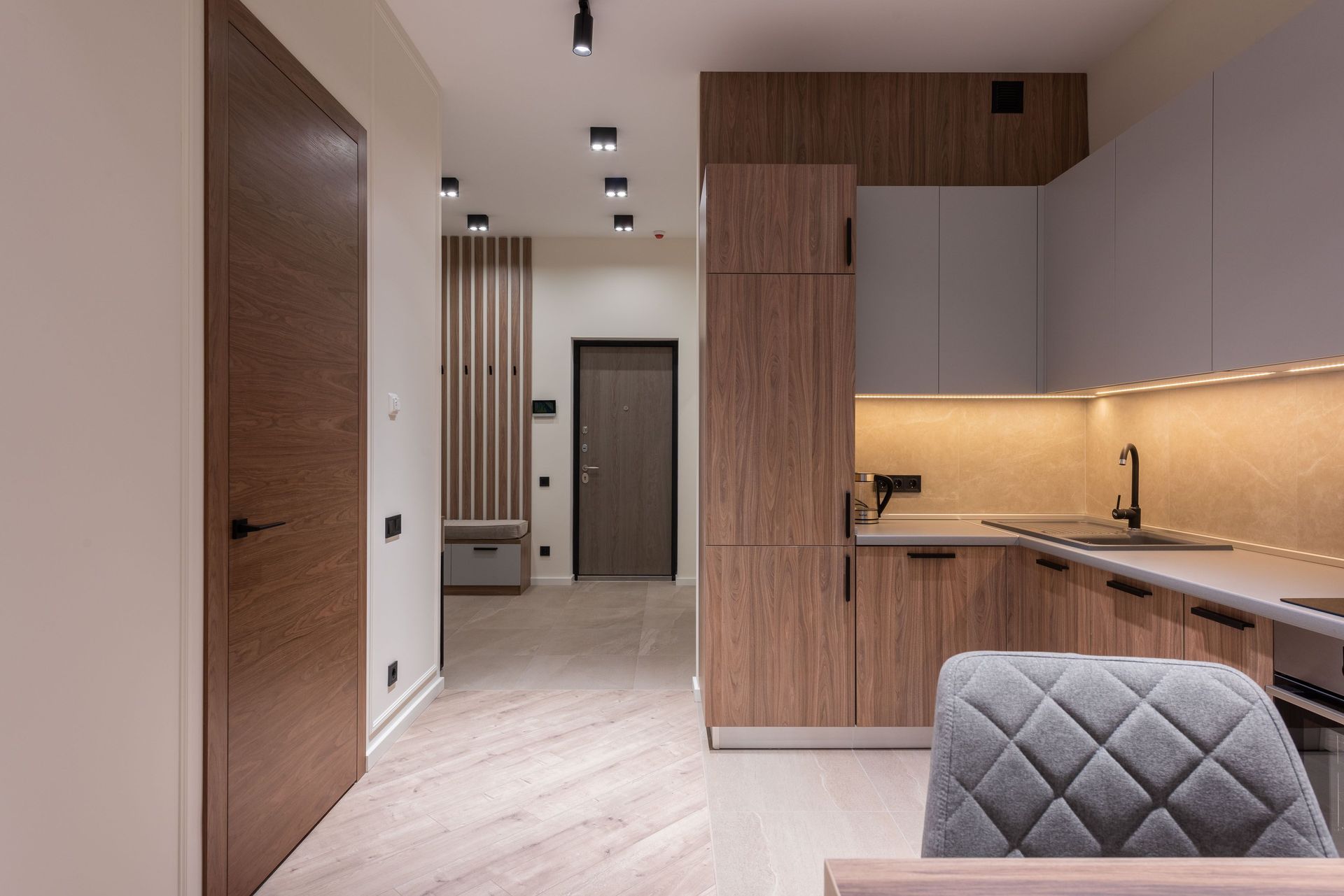 Modern minimalist kitchen and hallway with wood cabinets, light flooring, and warm under-cabinet lighting