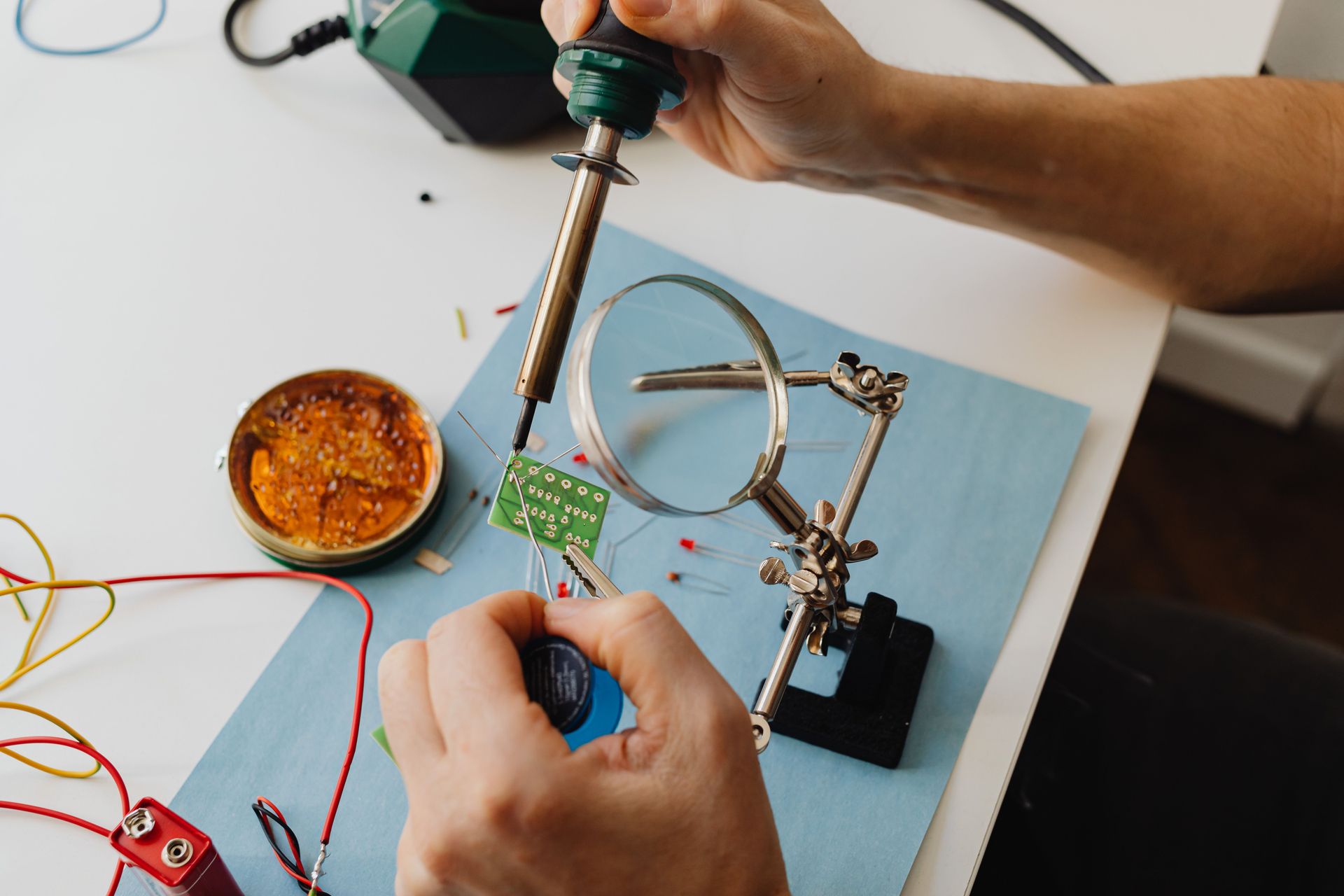 Hands soldering a green circuit board on a workbench with a soldering iron and helping hands tool