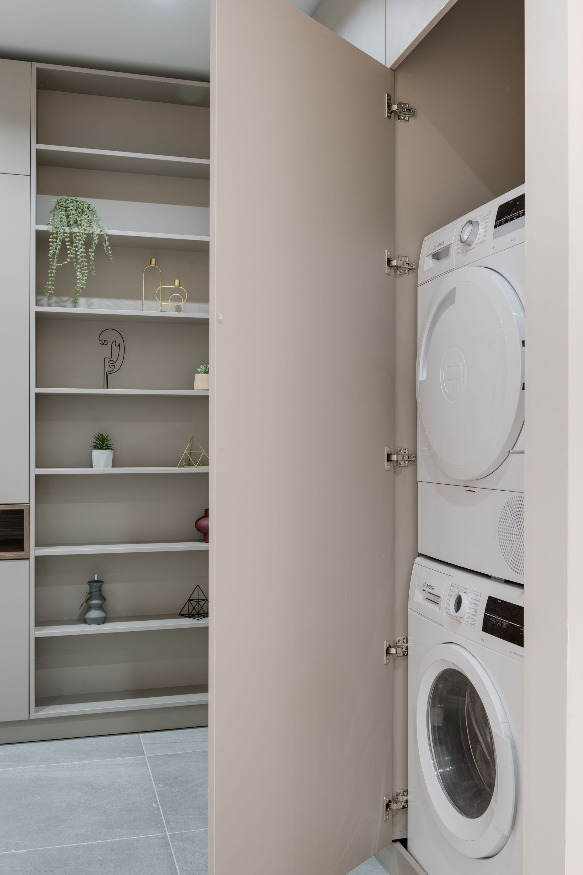 Laundry closet with stacked washer and dryer beside open shelves and a potted plant.