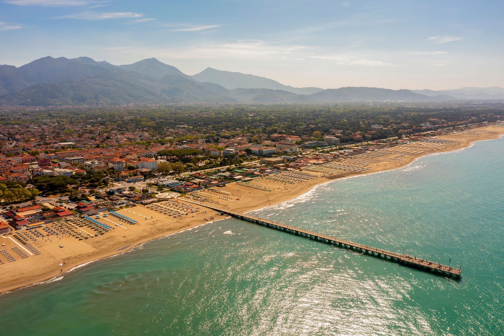Vista aerea di una città costiera con spiaggia sabbiosa, molo e montagne sullo sfondo in una giornata di sole.
