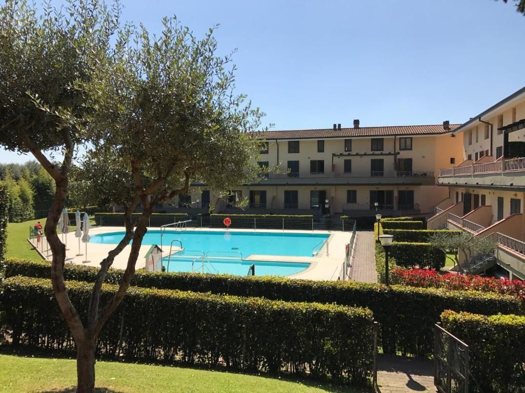 Piscina di fronte a un edificio a più piani con siepi verdi, alberi e cielo soleggiato.
