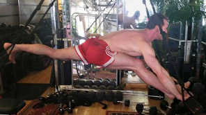A shirtless man in red shorts is stretching his legs in a gym