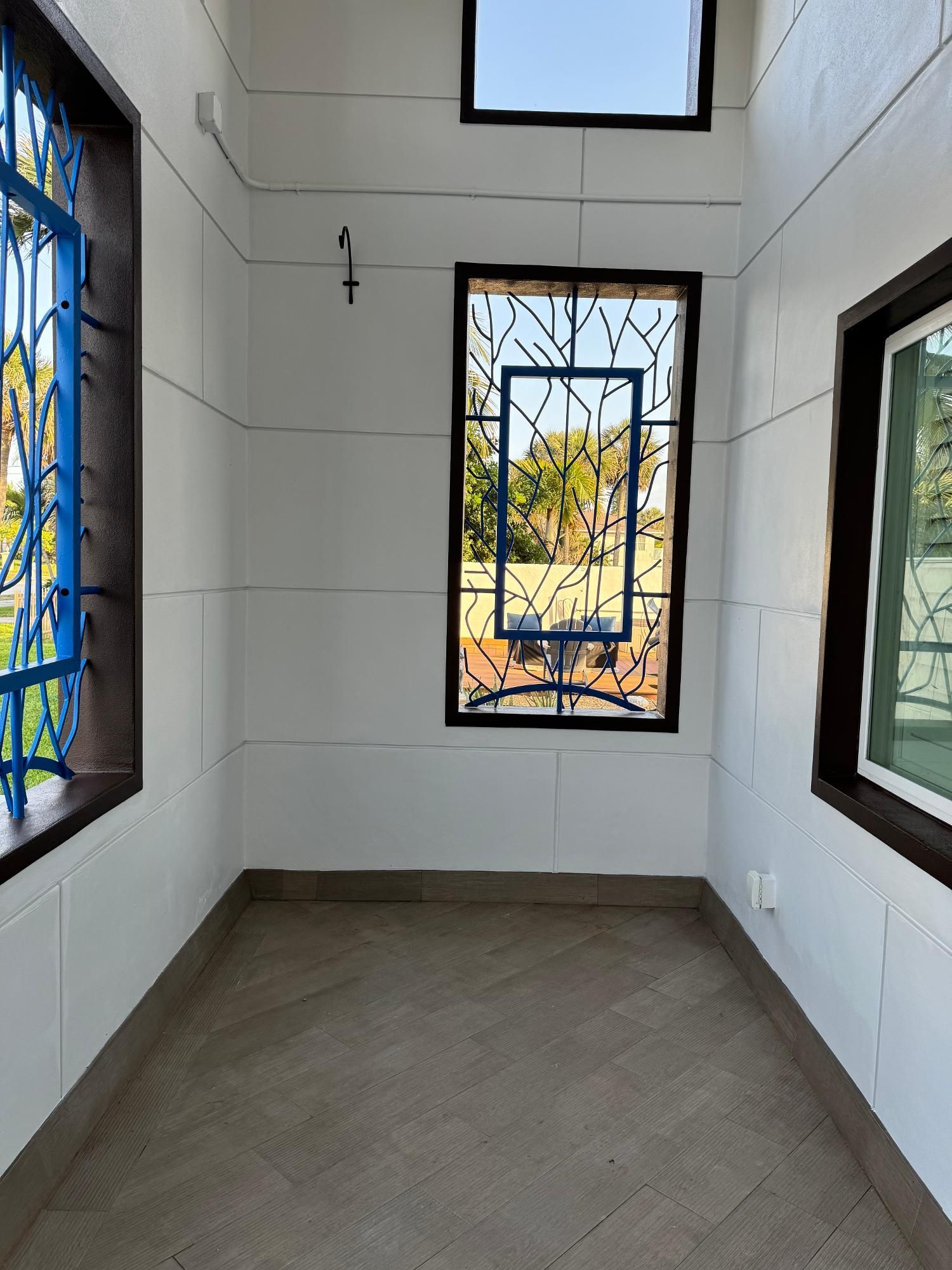 An empty, narrow sunroom with white paneled walls, tiled floor, and windows with blue decorative metal grilles.