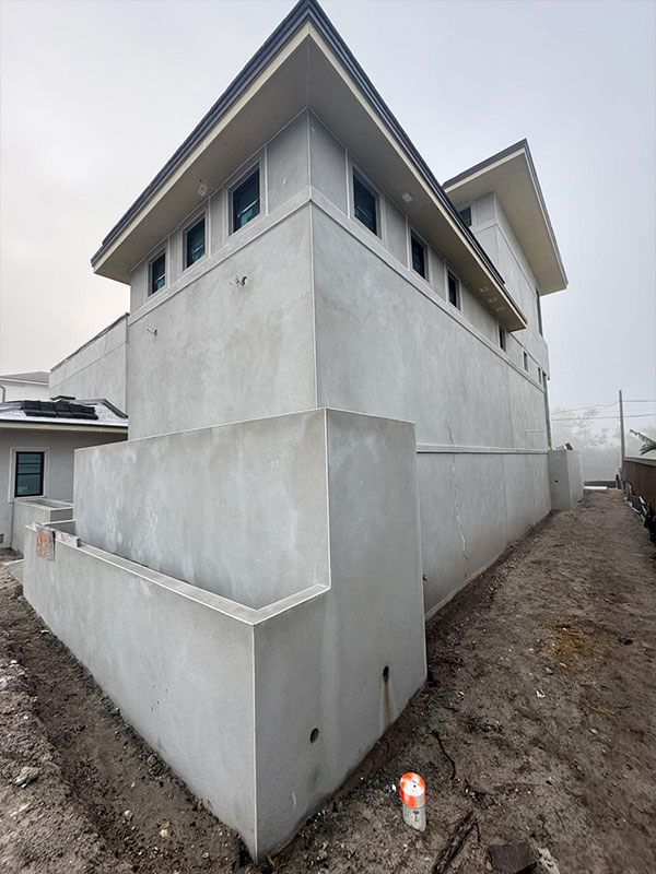 A multi-story building under construction with a smooth, gray stucco exterior and a row of high windows on a cloudy day.