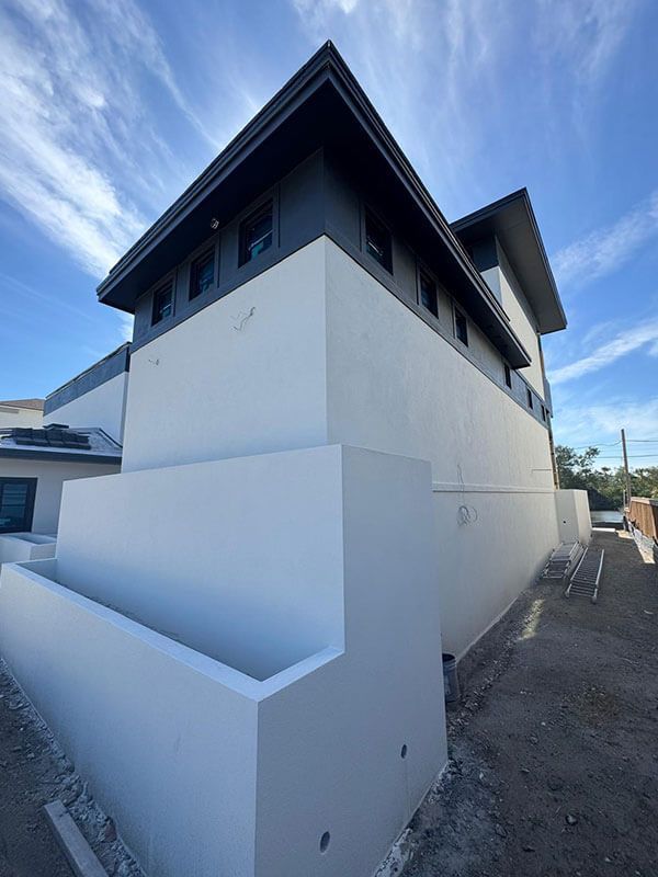 A modern white two-story building corner with dark gray trim under a blue sky, beside a narrow gravel walkway.